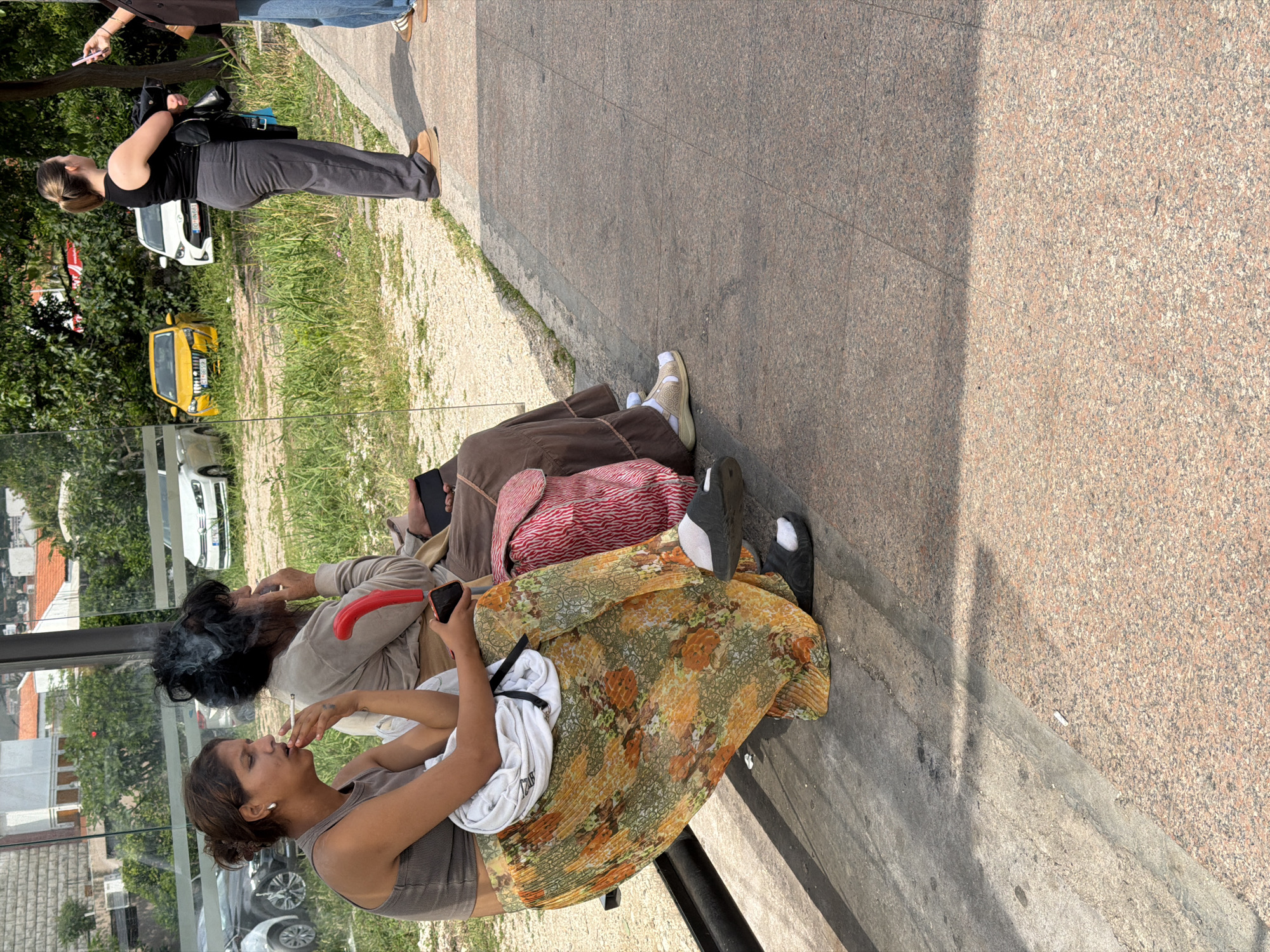 The image depicts a scene at a bus stop in Budva, Montenegro. Three women are seated on the bench. The woman closest to the camera is fair-skinned, wearing a grey tank top and a patterned yellow and orange skirt, and smoking a cigarette while holding a phone. Next to her is a person with dark, unkempt hair, smoking and holding a red cane. Behind them is another person wearing a brown shawl and tan footwear. The bus stop is covered in glass. Outside the bus stop, on a grass strip, are several cars, including a yellow vehicle. In the background, there are buildings and trees, indicative of an urban environment. Another woman stands nearby, holding a phone and a camera or bag. The area around the bus stop appears to be paved with stone.