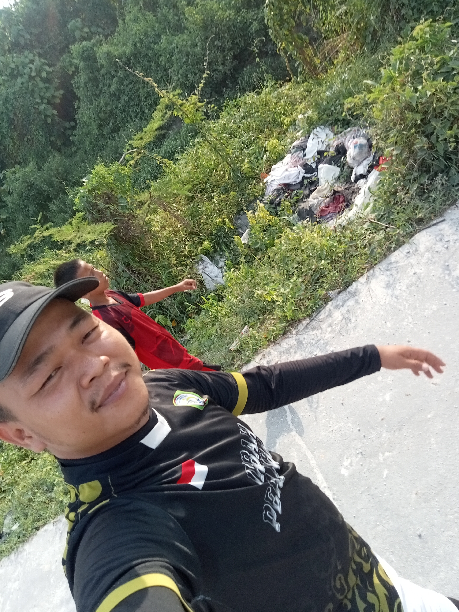 A man in a black shirt and hat is in the foreground, taking a selfie. Behind him, a younger person in a red shirt is standing with their arm extended. They are positioned on a paved surface next to a grassy area with a pile of discarded clothing. The background consists of dense green foliage. The scene appears to be outdoors, likely in a rural or semi-rural setting within Teluk Kenidai, Indonesia. The lighting suggests it is daytime, with sunlight illuminating the area.