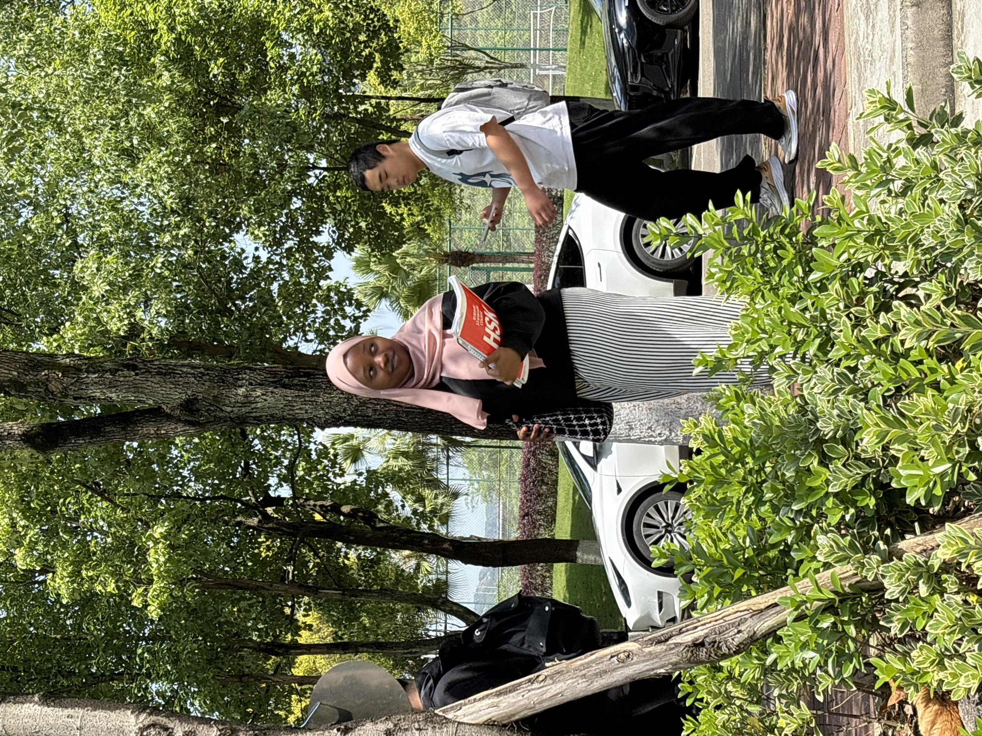 Two people stand outside on a paved area next to a road. A woman wearing a pink hijab, a black jacket, and a black and white striped skirt holds a red book. A man wearing a white t-shirt, black pants, and a light gray backpack looks at his phone. A tree partially obscures the subjects. Two cars, one white and one black, are parked behind the subjects. In the background, there is a fenced area with trees and water visible beyond. The location is the city of Jiangjin in China.