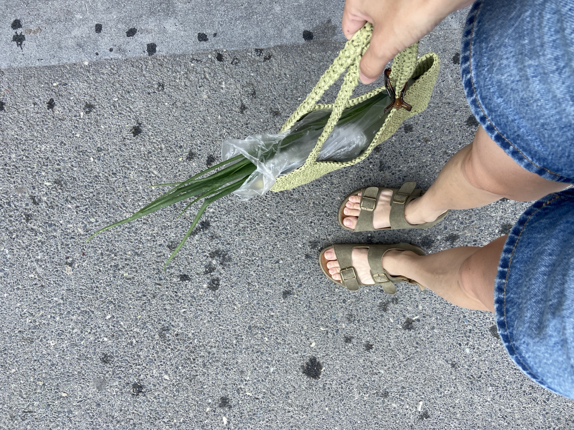The image shows a person's legs and feet wearing olive-green Birkenstock-style sandals and a denim skirt, standing on a gray asphalt surface. The person is holding a light green woven tote bag containing a bunch of green onions wrapped in plastic. The asphalt has scattered dark spots and small pebbles. The scene appears to be outdoors, likely on a sidewalk or street in Tyumen, Russia, given the context.