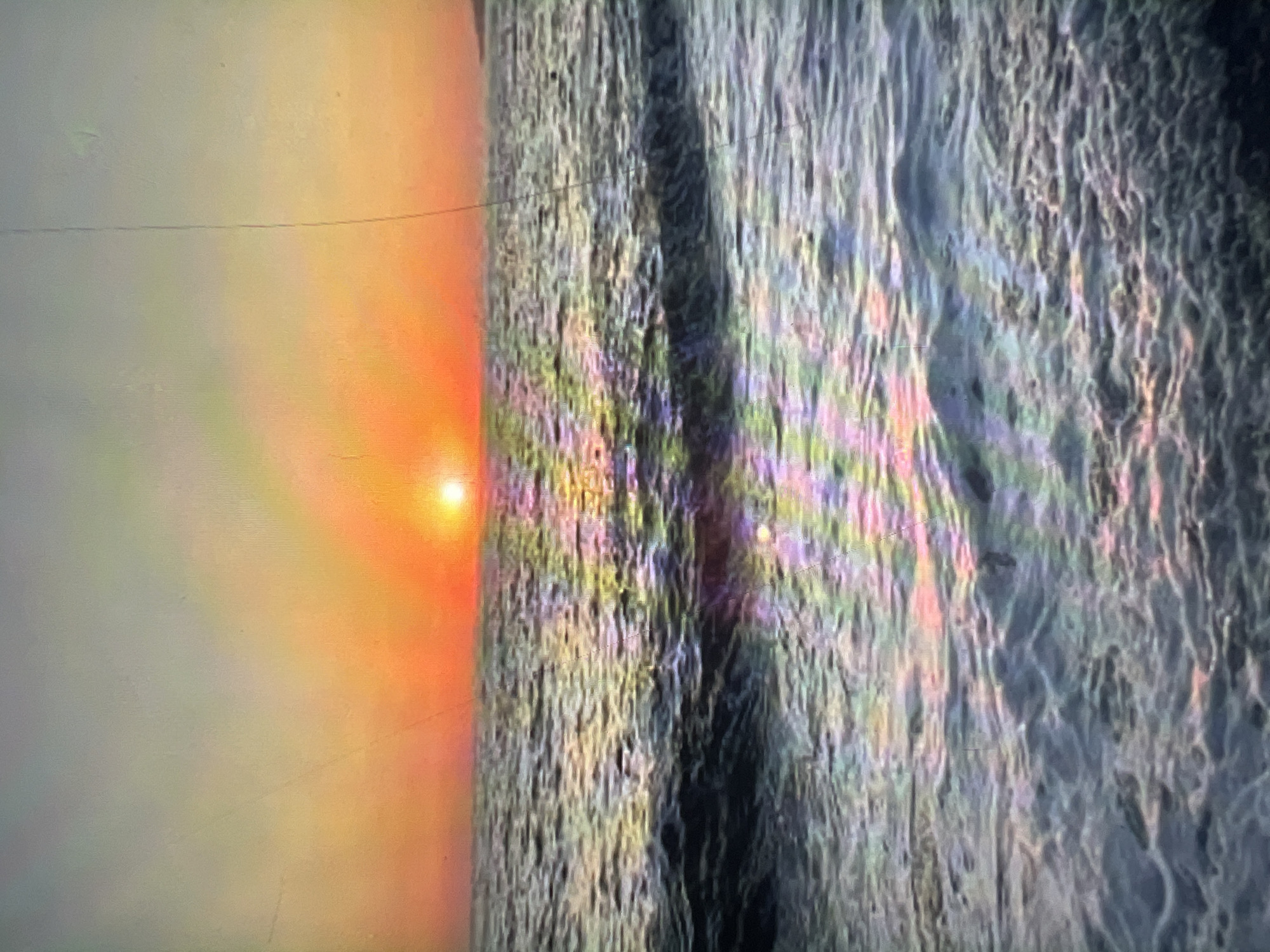 This media file depicts a sunset over the ocean, viewed from an abstract perspective. The sun is a bright, orange orb positioned on the left side of the image, casting a warm glow across the scene. The light from the sun diffuses into a soft haze, transitioning through shades of orange and yellow before fading into a pale, almost white sky.
The ocean occupies the right side of the frame, with its surface rendered in a textured, almost painterly style. The waves are not clearly defined but rather appear as a mosaic of colors, predominantly blues, grays, and greens, with streaks of pink and purple suggesting a prismatic effect from the sunlight reflecting on the water. A dark, almost black line runs vertically through the middle of the ocean, possibly representing a shadow or a deep trough in the water.
The overall impression is one of serene beauty and natural spectacle. The time of day is clearly sunset, indicated by the low angle of the sun and the warm hues. The weather appears calm, with the gentle undulation of the water suggesting minimal wind. No specific location cues are visible, and there is no text present in the image. The abstract rendering of the water might evoke a sense of awe and wonder at the interplay of light and water.