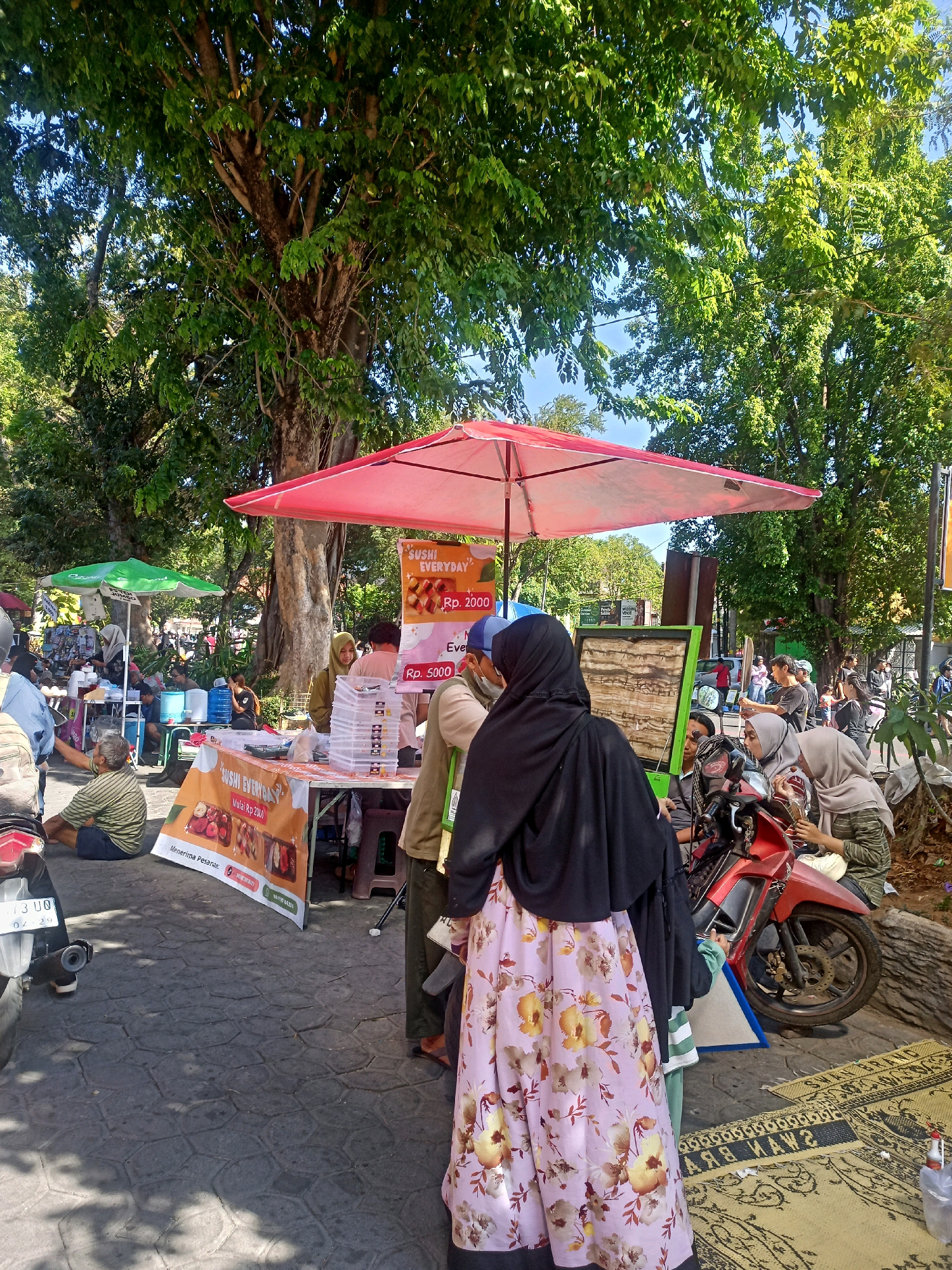 In Surakarta, Indonesia, a bustling outdoor market scene unfolds under a large, leafy tree. A prominent red umbrella shields a stall displaying sushi, with prices marked as Rp. 2000 and Rp. 5000. Several people are gathered around the stall, some examining the merchandise, while others appear to be engaged in conversation. A red motorcycle is parked nearby, adding to the lively atmosphere. The ground is paved with interlocking stones, and in the foreground, a woven mat lies on the ground. The image captures a slice of everyday life in a vibrant Indonesian city, with a focus on the commercial activity and social interactions taking place in the open air.