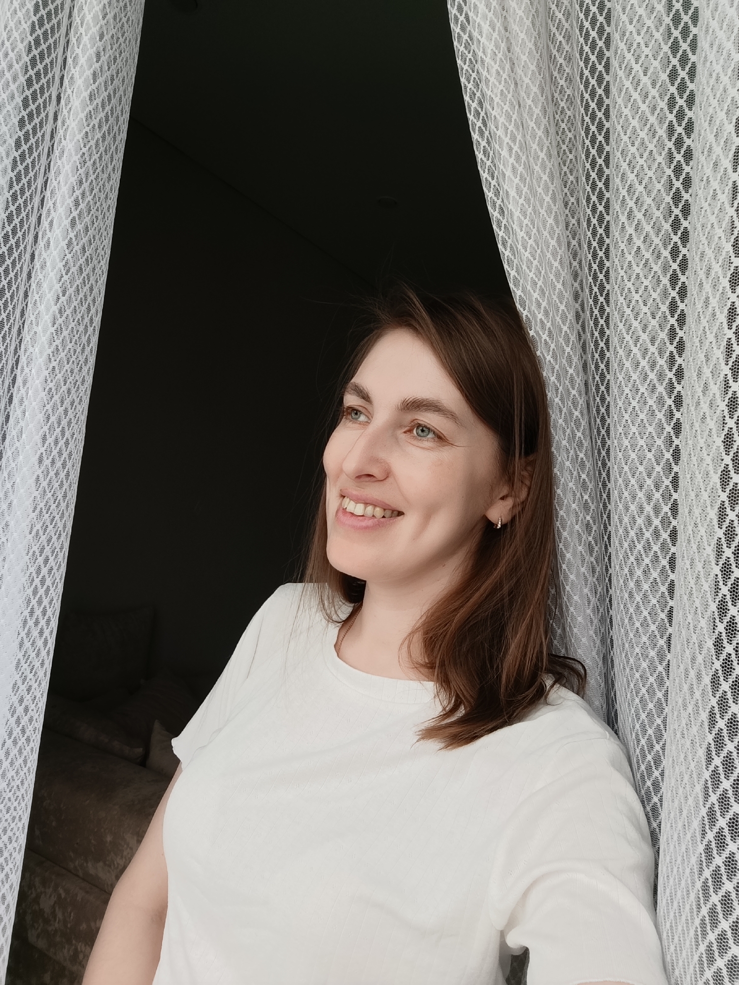 The image shows a woman with brown hair and a white t-shirt looking out of a window. The background is dark, but a couch can be seen in the back. White curtains frame the view. There is no information in the image that specifically states the location is in Chaikovsky, Russia.