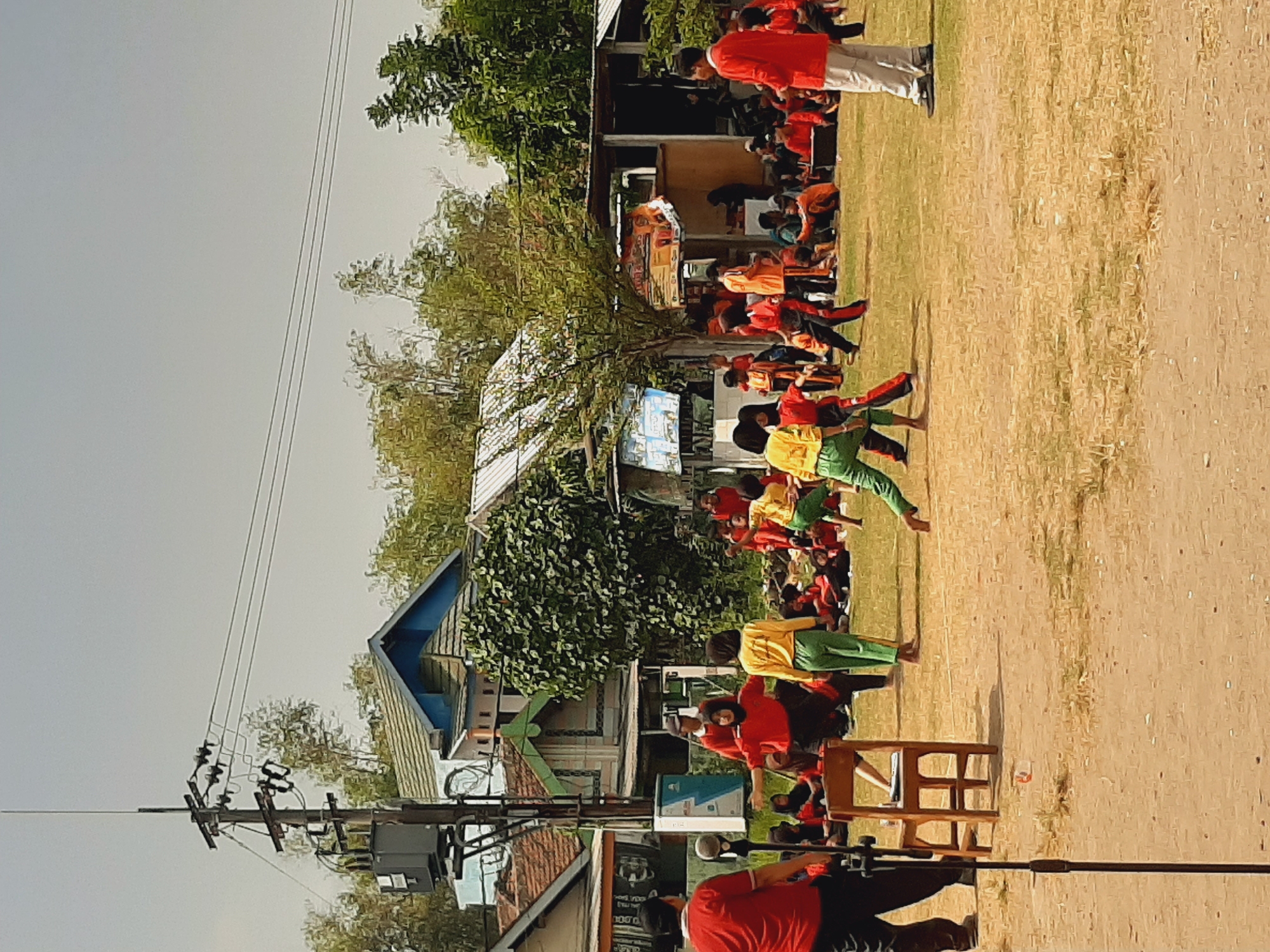 In Sumberjo, Indonesia, children are engaged in an outdoor activity on a dirt and grass field. A group of children, dressed in a mix of red and green uniforms, are participating in what appears to be a game or competition. One child in a yellow shirt and green pants is in motion, facing away from the camera, while another child in similar attire is to their left. In the background, a larger group of spectators, many wearing red, are seated or standing. A wooden chair is positioned near the center foreground, with a microphone stand to its left. The environment includes several buildings with corrugated metal roofs, trees, and a utility pole with electrical equipment. The sky is overcast.