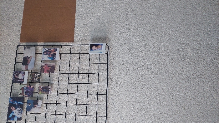 An indoor wall, likely in Santa María Ixtulco, Mexico, is visible, covered in a textured, light-colored finish, possibly stucco or rough plaster. Affixed to the upper left portion of this wall is a black metal grid-style photo display board, measuring approximately 12 squares wide by 12 squares high. Ten small, square-format photographs are attached to the upper section of this grid. These photographs depict various individuals and groups in different settings: a young male, groups of people outdoors, a smiling young female, two individuals embracing, a female with a child, two standing individuals, a group in red shirts, two individuals (one with blue hair), and a close-up of a female's face. To the right of the photo grid, a rectangular brown board, possibly cork, cardboard, or fiberboard, is mounted vertically on the wall. The wall beneath the grid and brown board is empty.