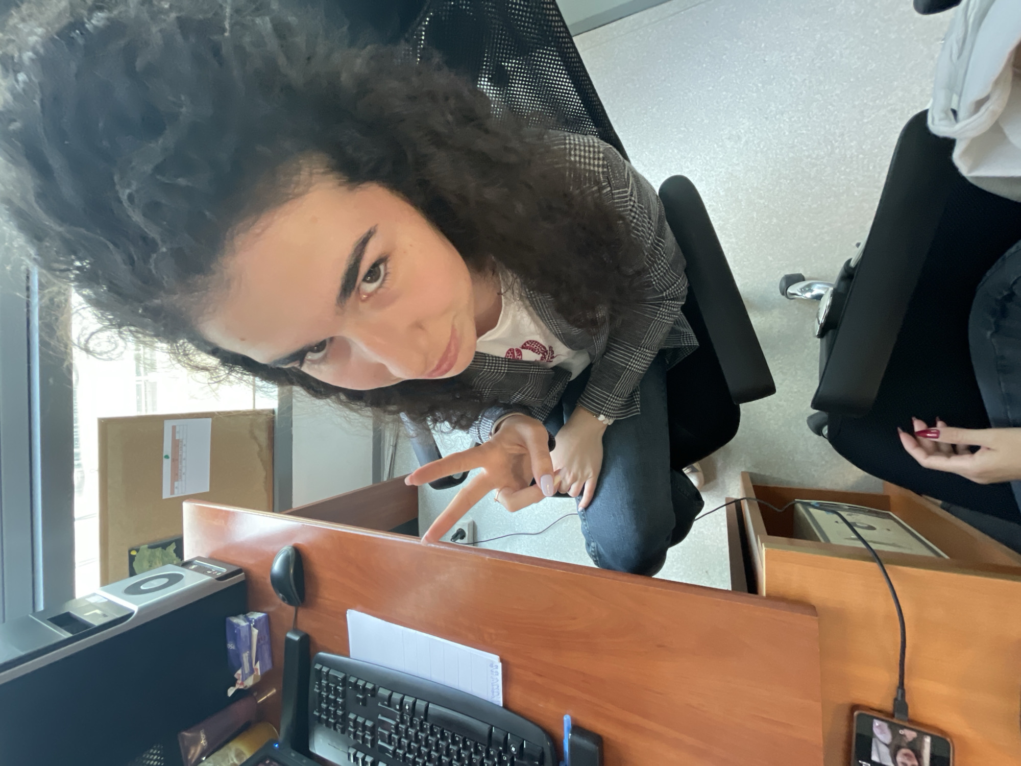 A young woman with curly dark hair is sitting in an office chair at a desk. She is wearing a gray plaid blazer over a white t-shirt and blue jeans. She is making a peace sign with her right hand. On the desk, there is a computer monitor, keyboard, mouse, a stack of white paper, and various office supplies. The desk is made of light brown wood. To the left of the monitor, there is a cardboard box. Another chair is visible to the right of the woman, with another person partially visible sitting there. The background shows a tiled floor and what appears to be an office environment. A phone plugged in is visible in the bottom right corner of the image. It is not possible to determine the exact location in Sofia, Bulgaria without more information.