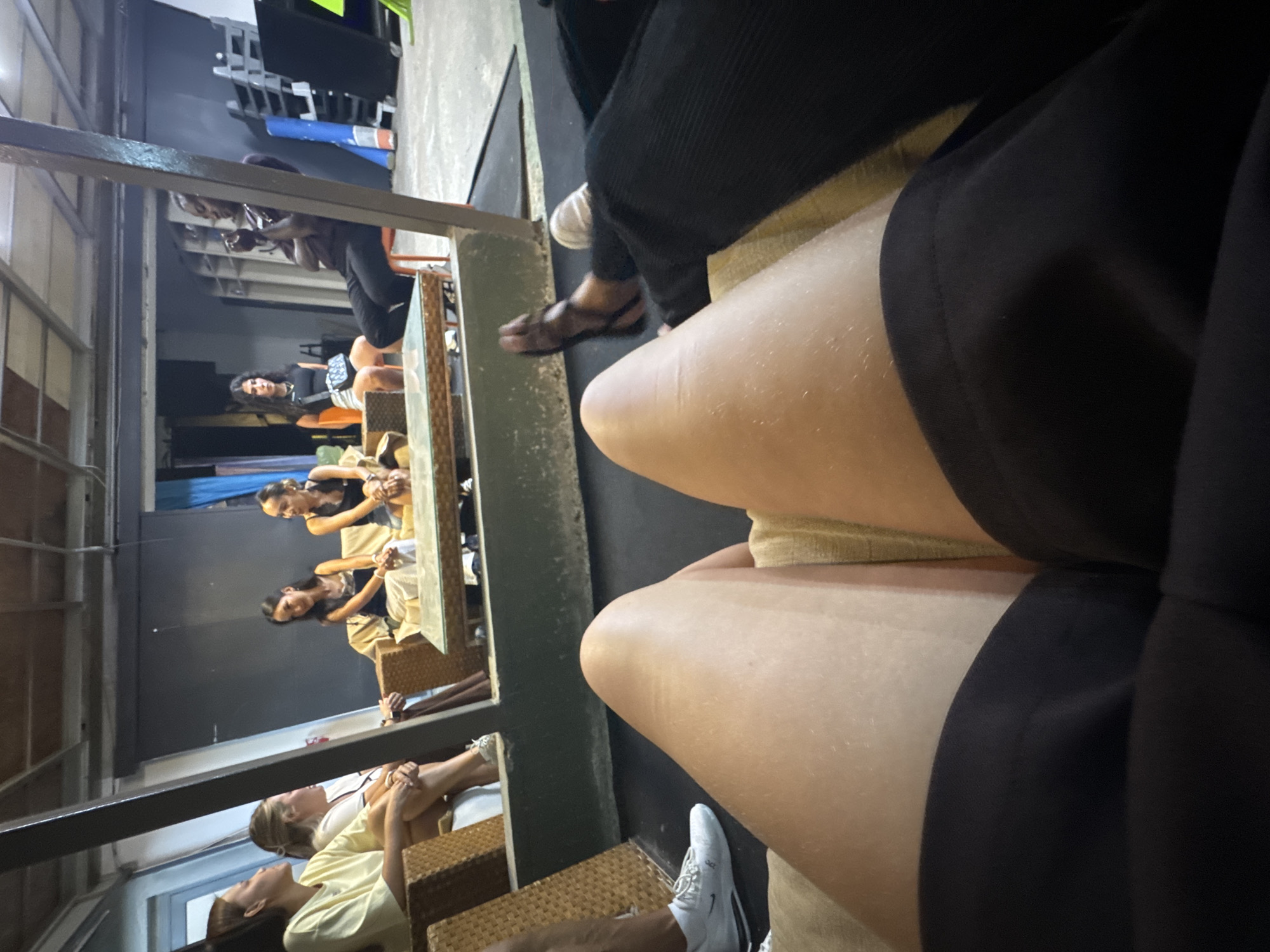 A first-person perspective shot shows legs in the foreground, with a group of people in the background. The scene is indoors, possibly a cafe or lounge, with a modern industrial aesthetic. The background features a table with several women sitting around it, engaged in conversation. One woman on the right is seated on a chair, while the others are on a couch. The environment includes exposed beams, concrete walls, and a few pieces of gym equipment visible in the distance, suggesting a multi-functional space. The lighting is dim, contributing to the relaxed atmosphere. The location is the city of Aksu, Turkey.