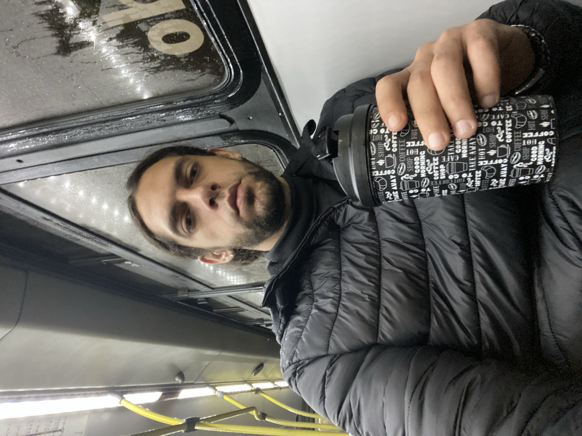 A man with medium-length brown hair and a beard is holding a coffee cup in his right hand. He is wearing a black puffer jacket. He appears to be on public transportation, possibly a bus, as indicated by the presence of handrails and the ceiling structure. There is a window with condensation or raindrops on it. The cup has a black and white coffee-themed design on it. It is not possible to determine the exact location in Buenos Aires from the image alone.