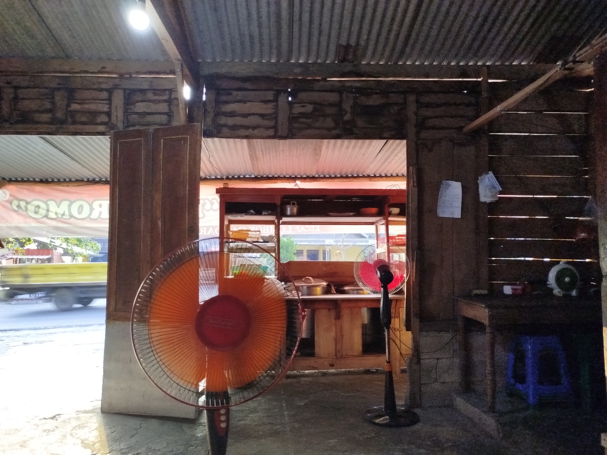 The image depicts the interior of a humble establishment, likely a food stall or small restaurant, situated in Pojok, Indonesia. The structure is rustic, with walls constructed from weathered wood planks and a corrugated metal roof, allowing natural light to filter through gaps. In the foreground, a large orange standing fan is prominently positioned. To its right, a smaller black standing fan with red blades is also visible.
The background reveals a counter or serving area with shelves displaying various items, including what appear to be containers and bowls. Beyond this, a window or opening offers a view of the outside, where a yellow truck is moving on a paved road under a bright sky. The ground inside the establishment is a concrete floor with an uneven texture. Two blue plastic stools are visible near a wooden table on the right side of the image, suggesting a seating area. Documents are pinned to the wooden wall on the right. The overall impression is one of a functional, open-air space designed for serving food in a tropical climate.