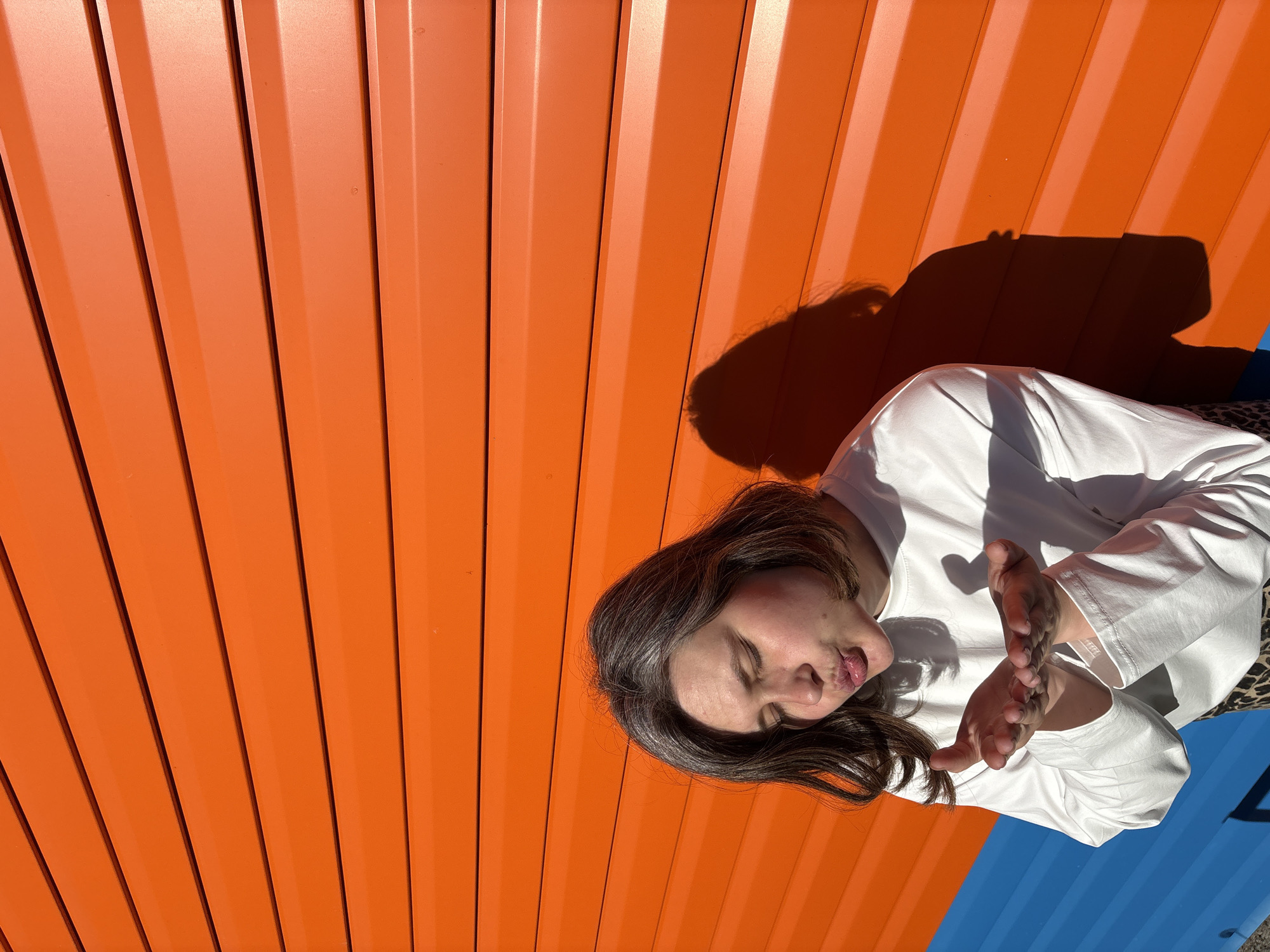A medium shot shows a light-skinned woman with dark hair wearing a white long-sleeved shirt. She is looking down with closed eyes and pursed lips, with her hands held up in front of her. The background is a bright orange corrugated metal wall on the left side, and a blue surface visible at the lower right corner. The photograph appears to be taken outside in bright sunlight, as indicated by the harsh shadows. The location is Chaikovsky, Russia.