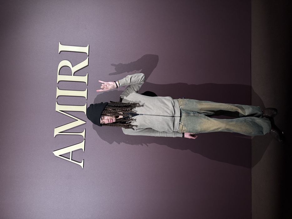 A single individual is depicted in a full-body portrait against a dark plum-colored wall. The setting is an indoor event space or retail environment associated with the Amiri fashion brand in Milan, Italy, as indicated by the large, metallic gold-colored "AMIRI" logo affixed to the wall above the individual.
The subject is an individual, appearing male, with light skin and dark, dreadlocked hair partially covered by a black knitted beanie. The individual wears a light beige or taupe full-zip track jacket over a dark undershirt with a visible pendant. The lower attire consists of loose-fitting, distressed denim jeans featuring a faded light blue and yellowish-brown wash. Dark brown or black boots with a slightly pointed toe cover the feet. Several dark beaded bracelets are worn on the left wrist.
The individual stands facing forward, with their head slightly turned, exhibiting a neutral or relaxed facial expression. Their right arm is raised, with the hand forming a "rock on" gesture (corna), while the left arm hangs relaxed at the side. Artificial lighting illuminates the scene from the front, casting a distinct, elongated shadow of the individual onto the wall to their right (viewer's left). The floor is a dark, uniform surface.