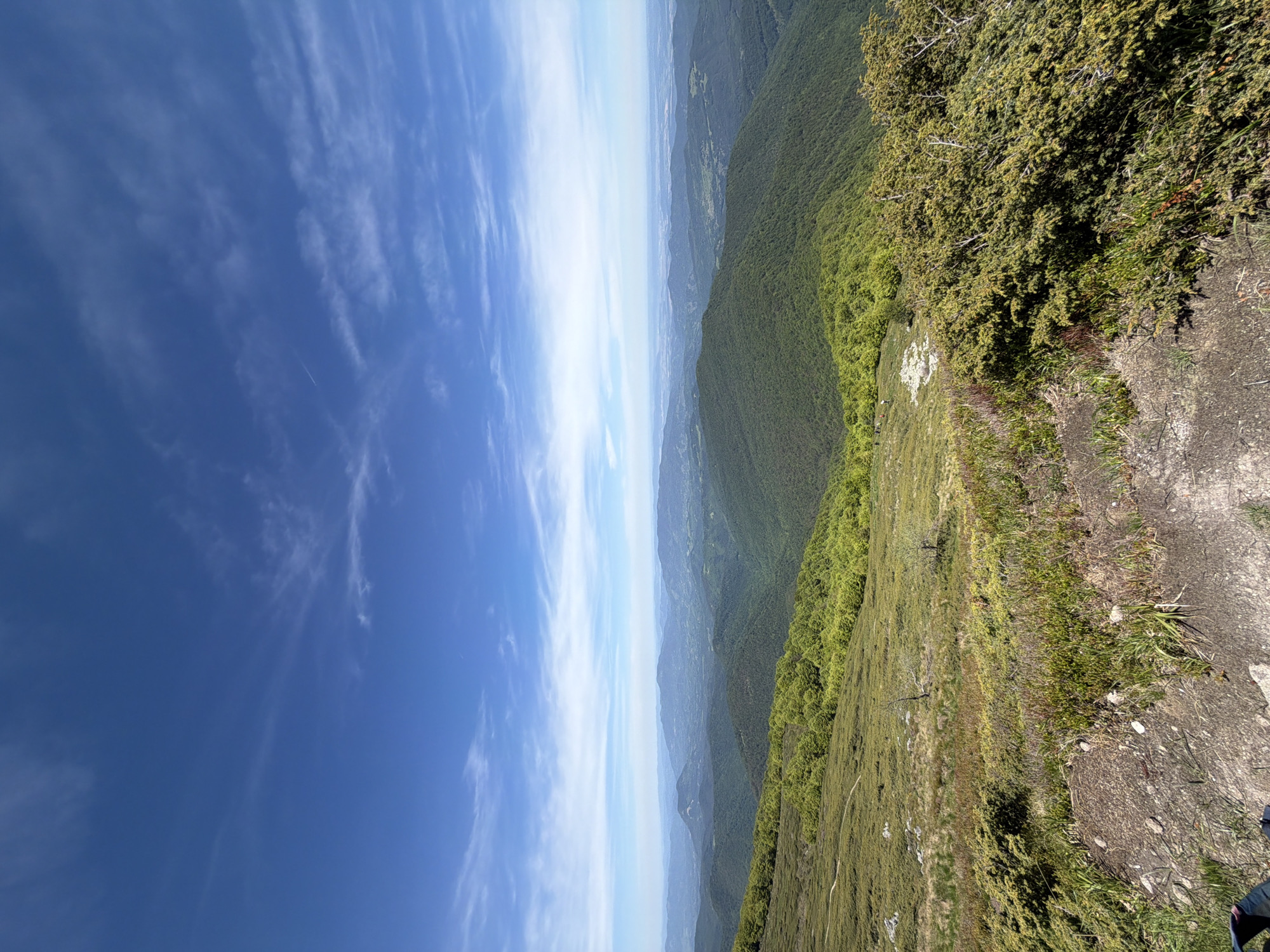The image depicts a landscape viewed from a high vantage point, possibly a mountain peak. The sky, a clear blue with wispy cirrus clouds, occupies the left half of the image. A series of forested mountain ranges stretch into the distance, gradually fading into a hazy horizon. The right side of the image shows the immediate foreground, covered with green vegetation and rocky terrain. The scene is likely located in a mountainous region. The specific city of Stokite, Bulgaria, cannot be discerned from the image itself. There are no visible people or subjects in this image.