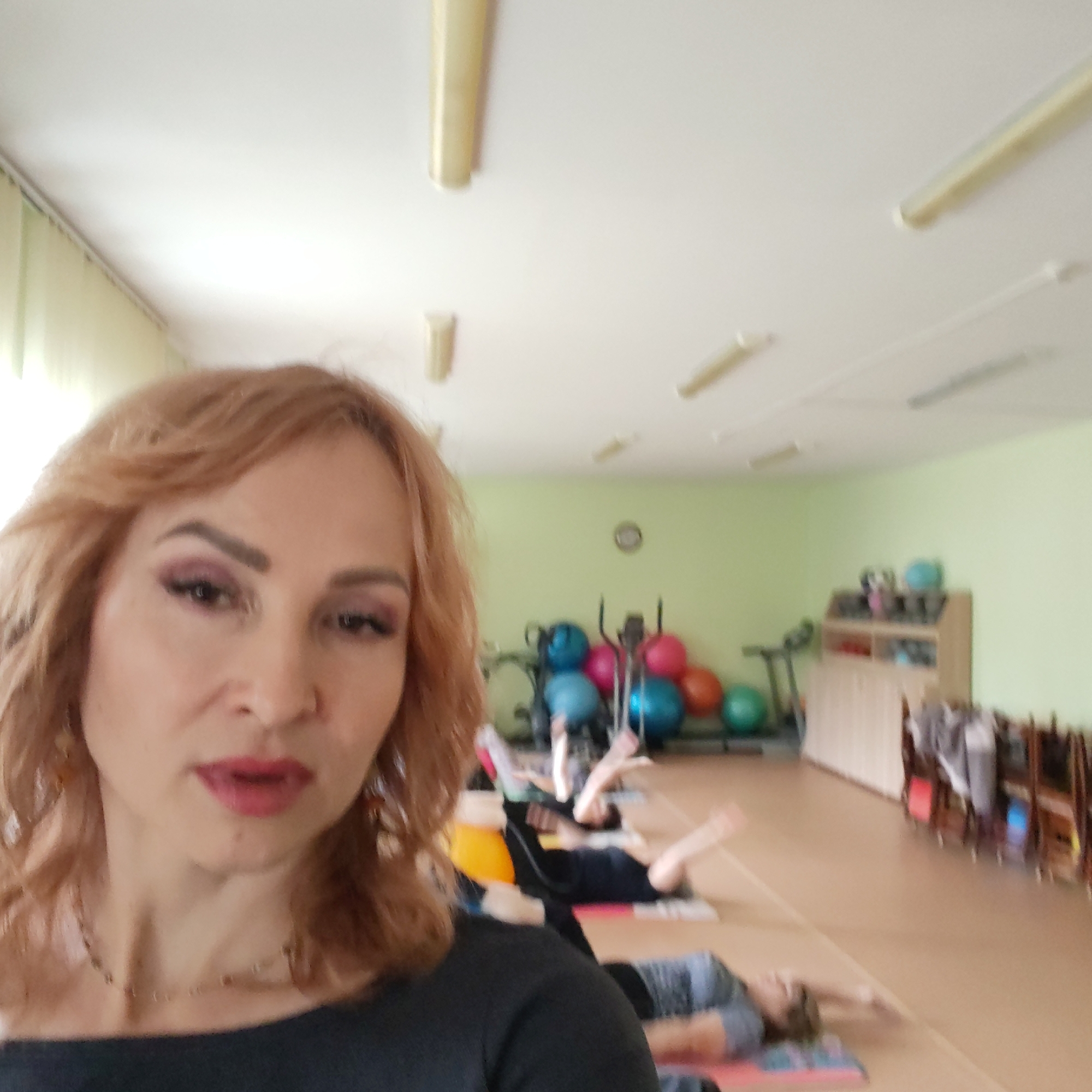 The image shows a woman taking a selfie in a room where a group of people is doing a yoga or stretching class. The woman has reddish-blonde hair, light skin, and is wearing makeup. She is wearing a dark-colored shirt. Behind her, a line of people is lying on yoga mats, stretching their arms overhead. Further back, there is exercise equipment including fitness balls and exercise machines. The room has light green walls, a light-colored floor, and fluorescent lighting on the ceiling. There is a shelf unit along the back wall. The location is in Chaikovsky, Russia.