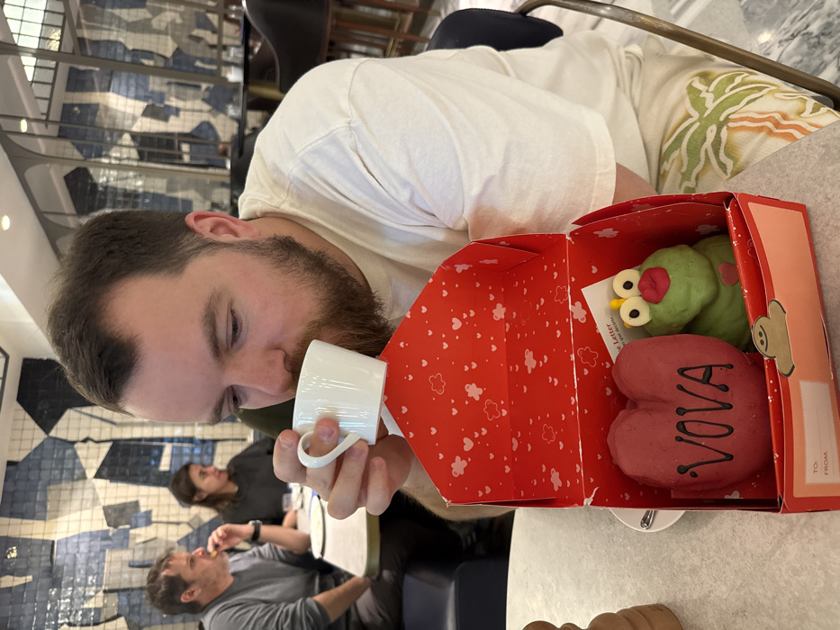 A bearded male, wearing a light-colored t-shirt and patterned shorts, is seated at a white table within a restaurant, identified by its distinctive blue and white geometric tiled walls and arched metal frames as likely El Nacional in Barcelona, Spain. He is shown bringing a small white ceramic cup to his mouth. Directly in front of him is an open red gift box, internally patterned with white hearts and clouds. Inside the box are two custom confectionery items: a dark pink heart-shaped piece inscribed with "VOVA" in black, and a green frog-like figure featuring large eyes, red lips, and a small pink heart on its chest. In the background, two other patrons are visible: a male in a grey t-shirt engaged in eating, and a female seated opposite him.