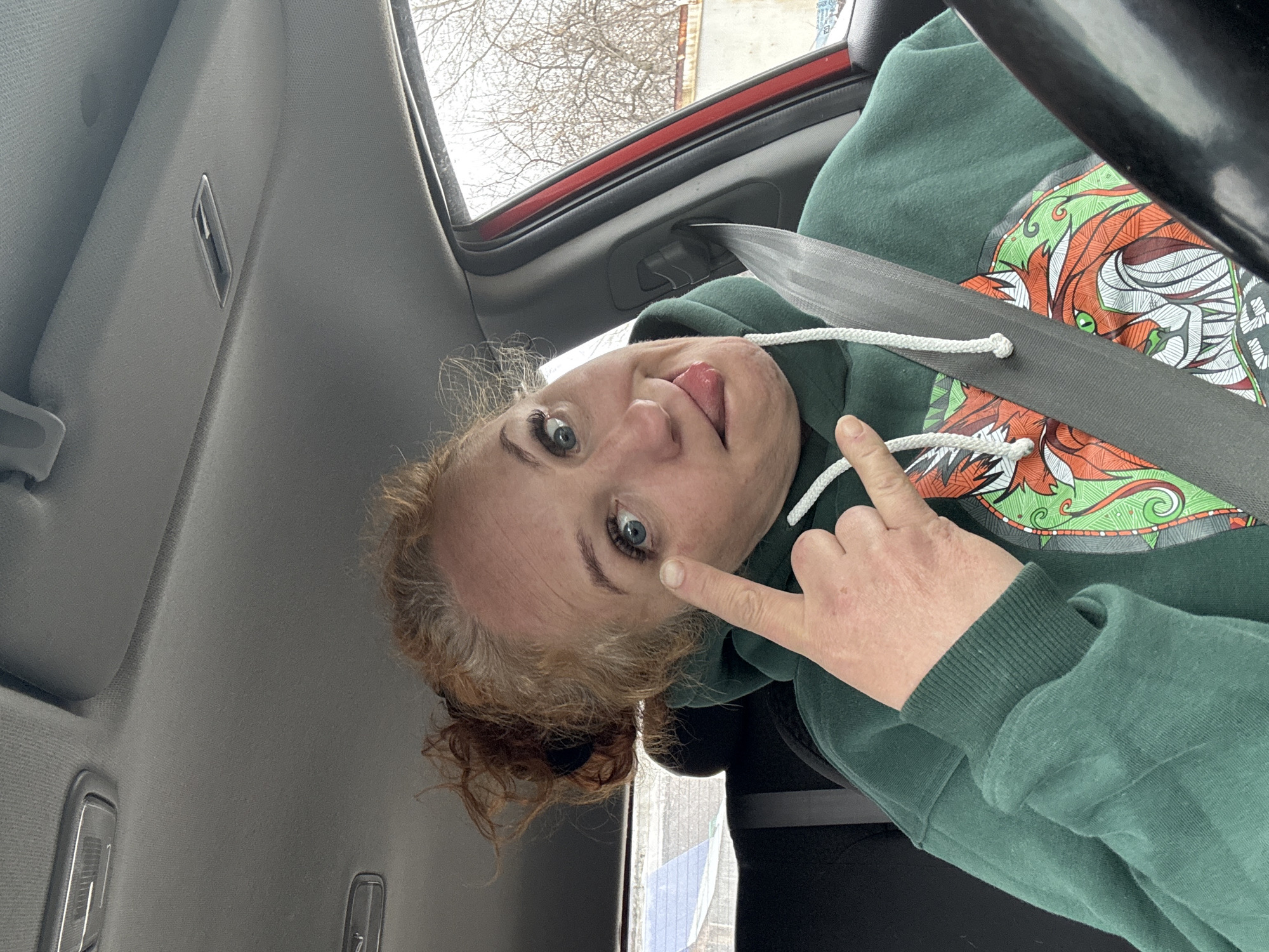 A woman is taking a selfie inside a vehicle. She has fair skin, red curly hair pulled back into a bun, and blue eyes. She is wearing a green hooded sweatshirt with a colorful design on the front and is wearing a seatbelt. Her finger is pointing towards her left eye, and her tongue is sticking out. The interior of the vehicle is gray, with the window showing a glimpse of trees and a building outside. The exact location in the city of Chaikovsky, Russia, cannot be determined from the visual data.