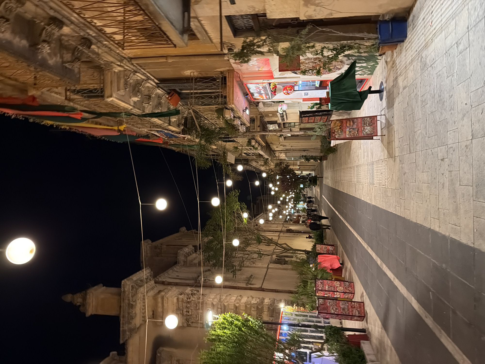 This image depicts a street scene at night in Ragusa, Italy. The street is lined with buildings, illuminated by hanging string lights. Red, yellow, and green flags are hung across the buildings. Several people are walking down the street. Along the right side of the street are food stands and displays. The street is paved with light-colored stones. A tall building is visible in the background on the left.