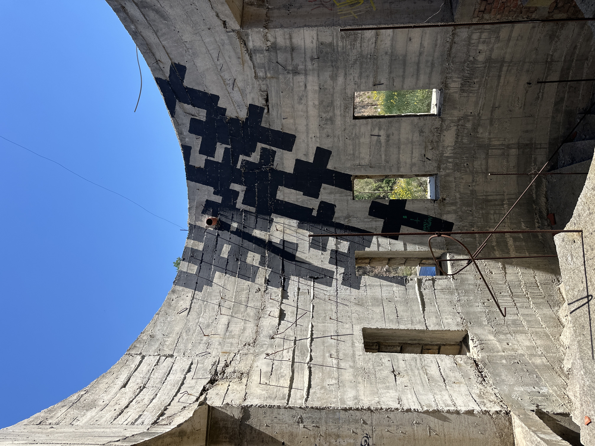 The image depicts the interior of a concrete structure located in Gornji Katun, Montenegro. The structure appears to be an abandoned building with exposed concrete walls and a curved archway. The walls feature numerous square or rectangular openings, potentially former windows, with glimpses of greenery visible through some. A pattern of large, black, cross-shaped graffiti is visible on the curved wall. Exposed rebar protrudes from the concrete in various places. The sky is visible through the archway.
