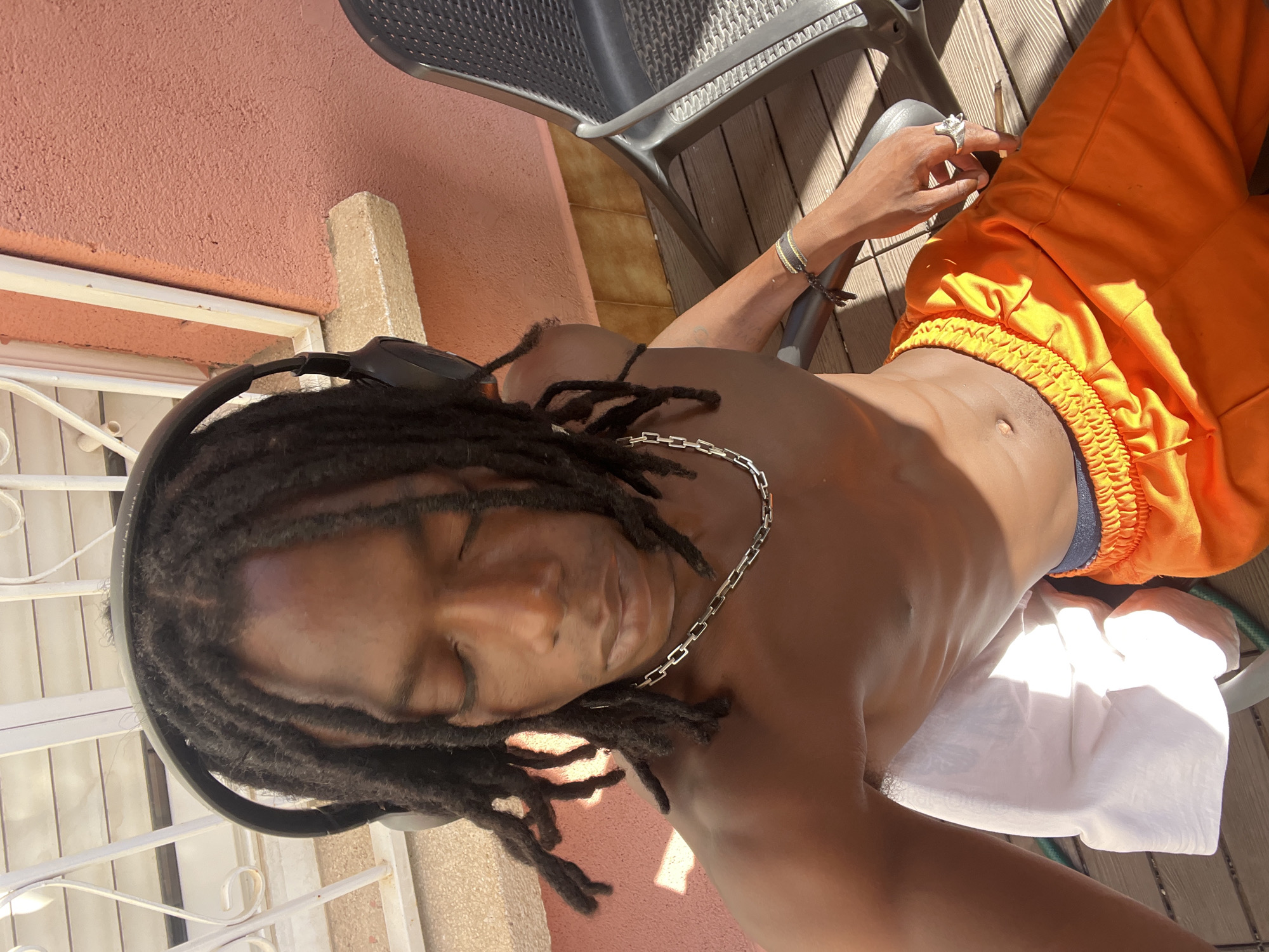 The media file shows a person with dreadlocks wearing headphones and a silver chain, lying on a white cloth outdoors. The person is wearing orange shorts and has rings on their fingers. Sunlight is casting shadows, suggesting it is daytime. The background includes a textured pink wall, a wooden deck, and a metal chair. A white metal railing is visible in the foreground. The overall impression is of a relaxed, sunny outdoor setting, possibly in a private courtyard or balcony. The orientation of the image is portrait, with the subject positioned centrally. The lighting and shadows indicate a bright, sunny day. The location is suggested to be Barcelona, Spain.