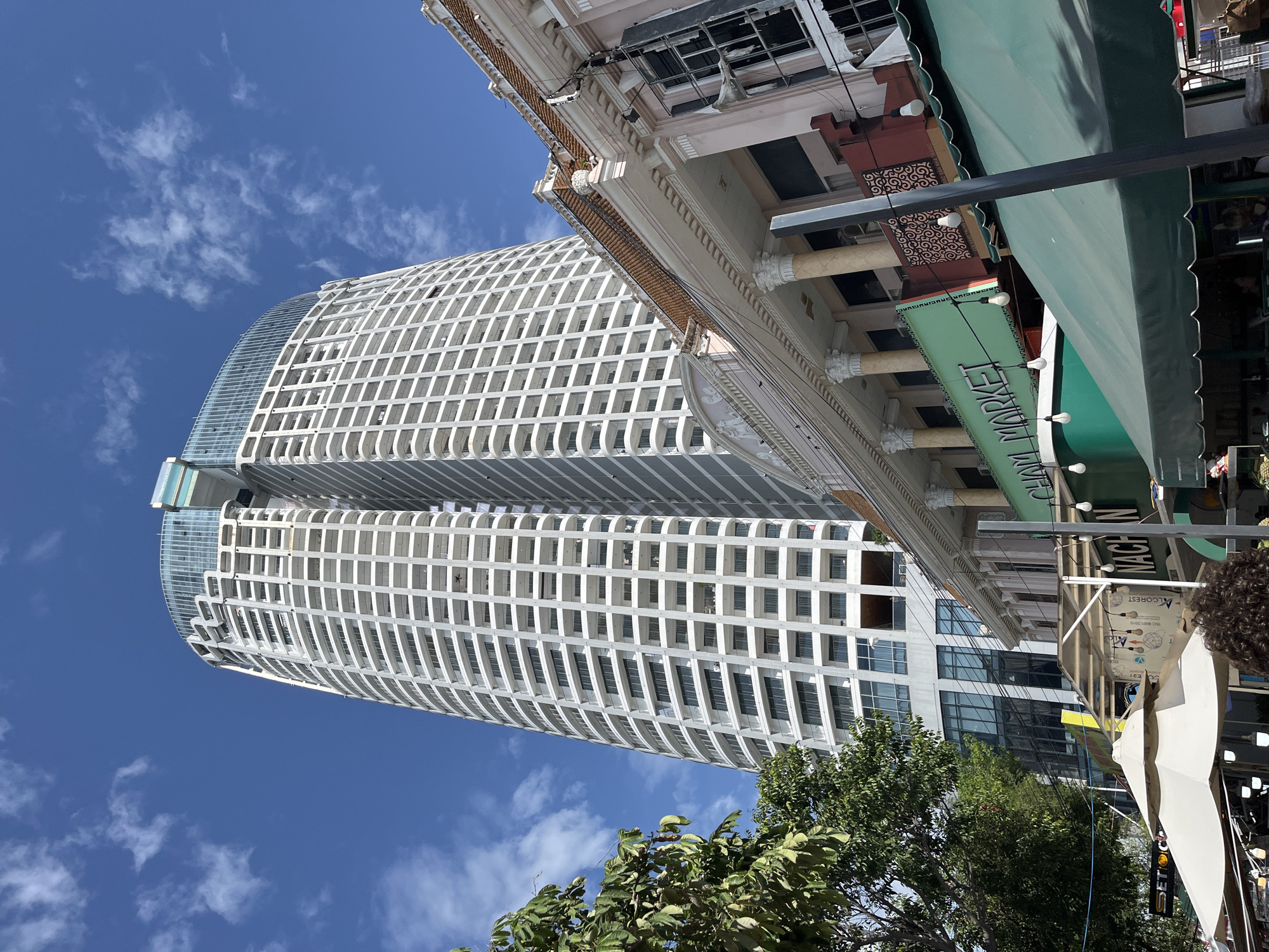 A tall, modern, white high-rise building with rounded corners and many balconies dominates the background of this urban scene in Nha Trang, Vietnam. The building's upper section is topped with a glass-enclosed structure. In the foreground, a row of older, pastel-colored buildings with ornate architectural details lines the street. A green awning stretches across the front of one of these buildings, displaying the illuminated text "CHÂM MARKET." Trees with lush green foliage are visible on the left side of the frame. The sky is a clear, vibrant blue with scattered white clouds. The overall impression is a juxtaposition of old and new architecture in a bustling city environment.