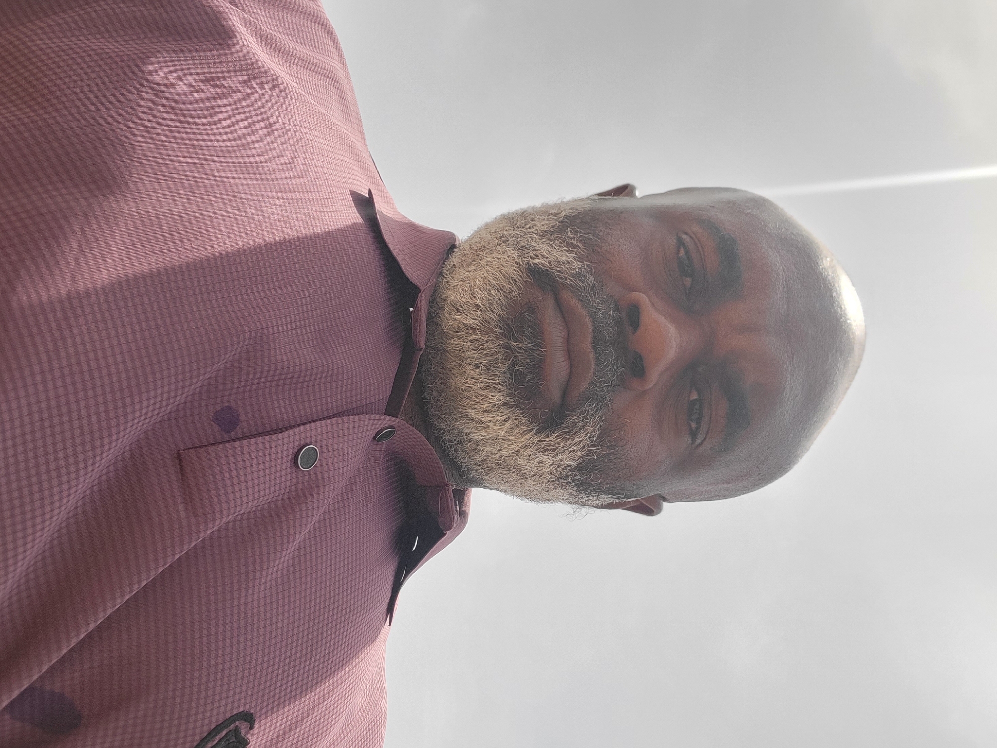 A close-up, low-angle shot shows a middle-aged, dark-skinned man with a bald head and a grey/white beard. He is wearing a maroon, collared shirt with a buttoned chest pocket. The sky is overcast and appears bright, filling the background. There is no indication of the location being Narayi, Nigeria in the image.