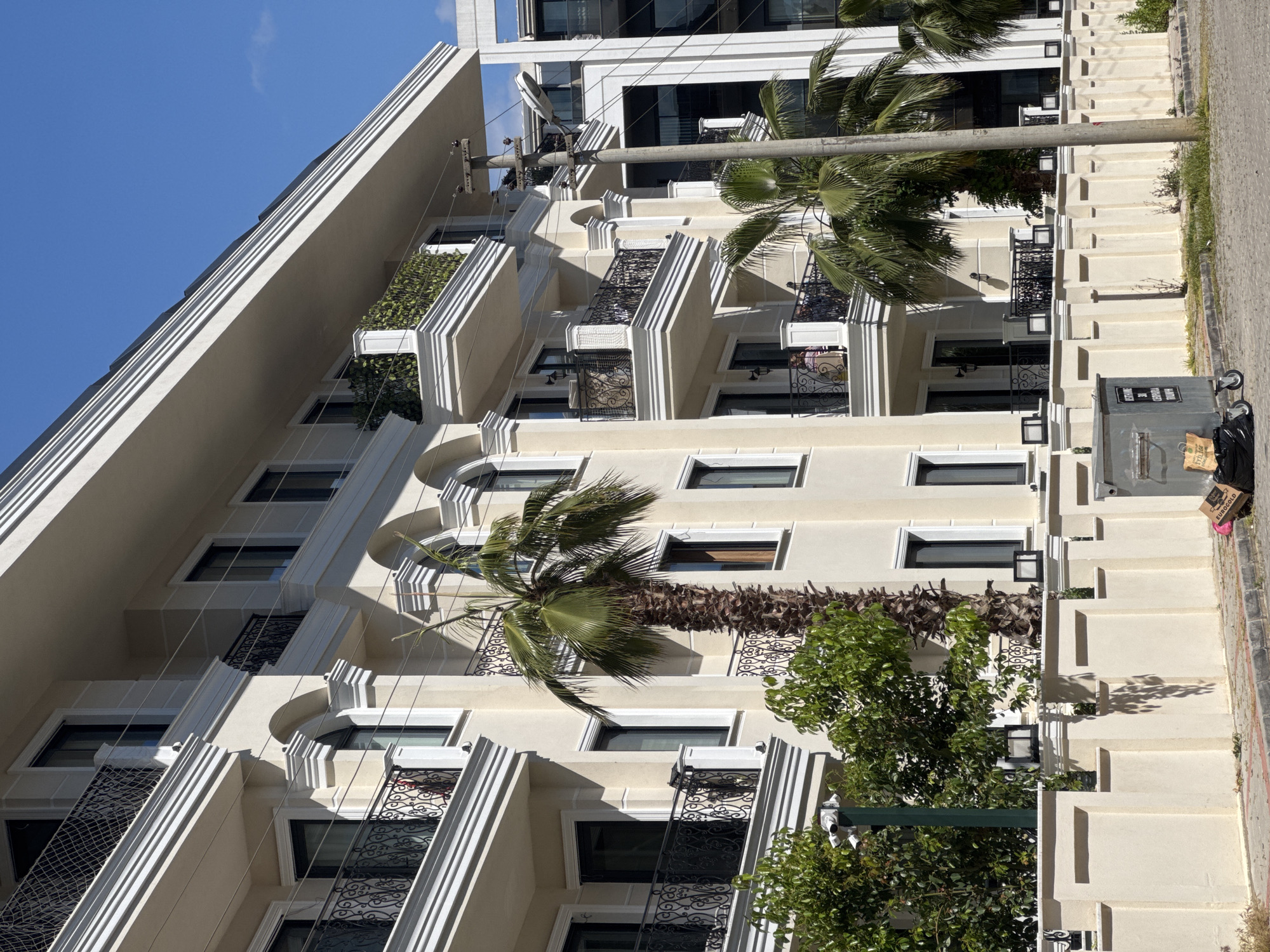 A multi-story building is shown. It has many balconies with wrought iron railings. Palm trees are in front of the building. A utility pole is also in front of the building. A trash receptacle is on the right side. The sky is blue. The location is Unknown, Unknown.