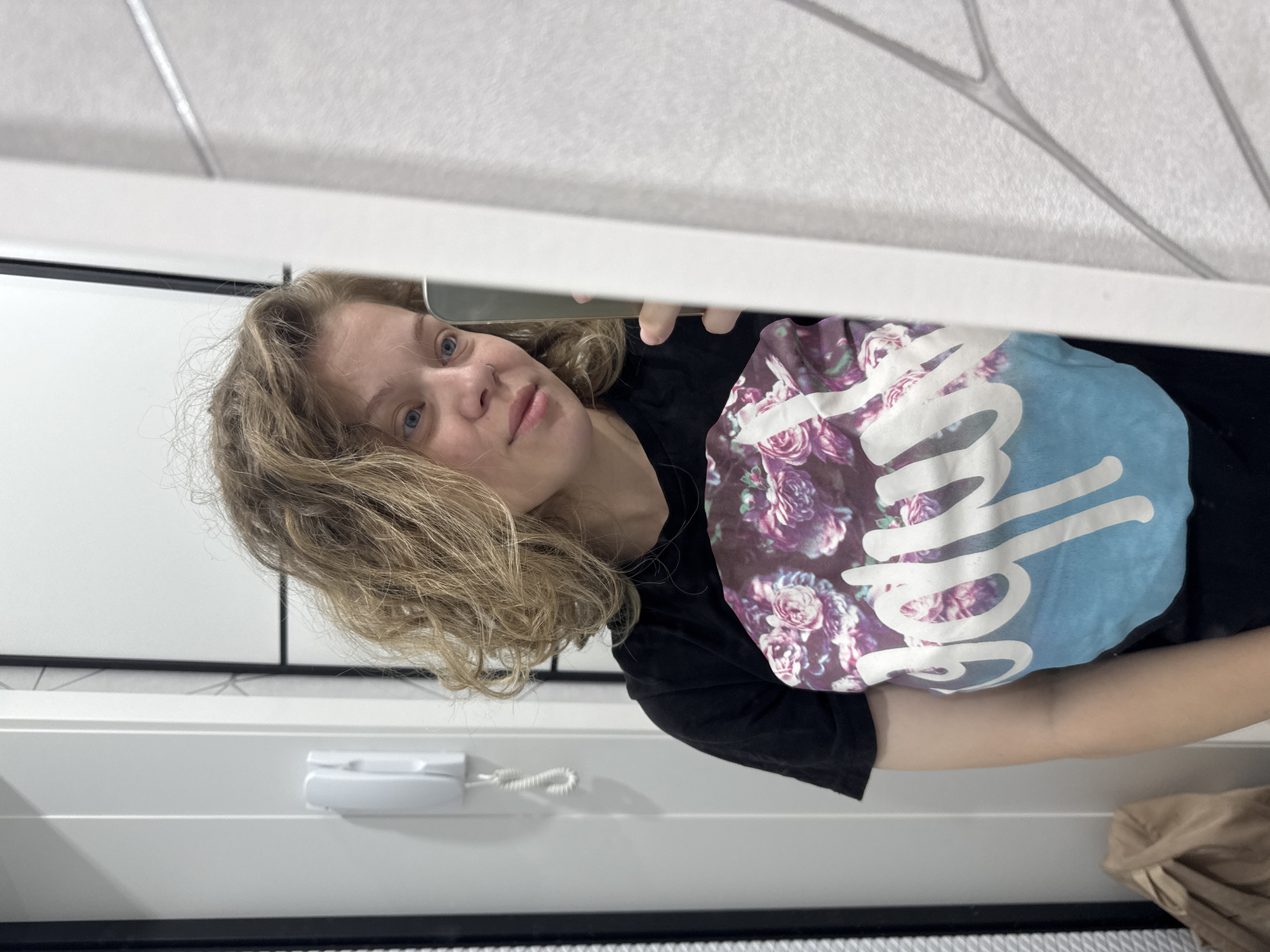 A young woman with light brown, curly hair takes a selfie in a mirror. She is wearing a black t-shirt with a floral print and white text. The background appears to be a home interior, possibly a bathroom or hallway, with a white wall and a door. A white intercom device is mounted on the wall to the left. A brown paper bag is visible on the floor to the left of the frame. The location is specified as Mikhaylovsk, Russia.