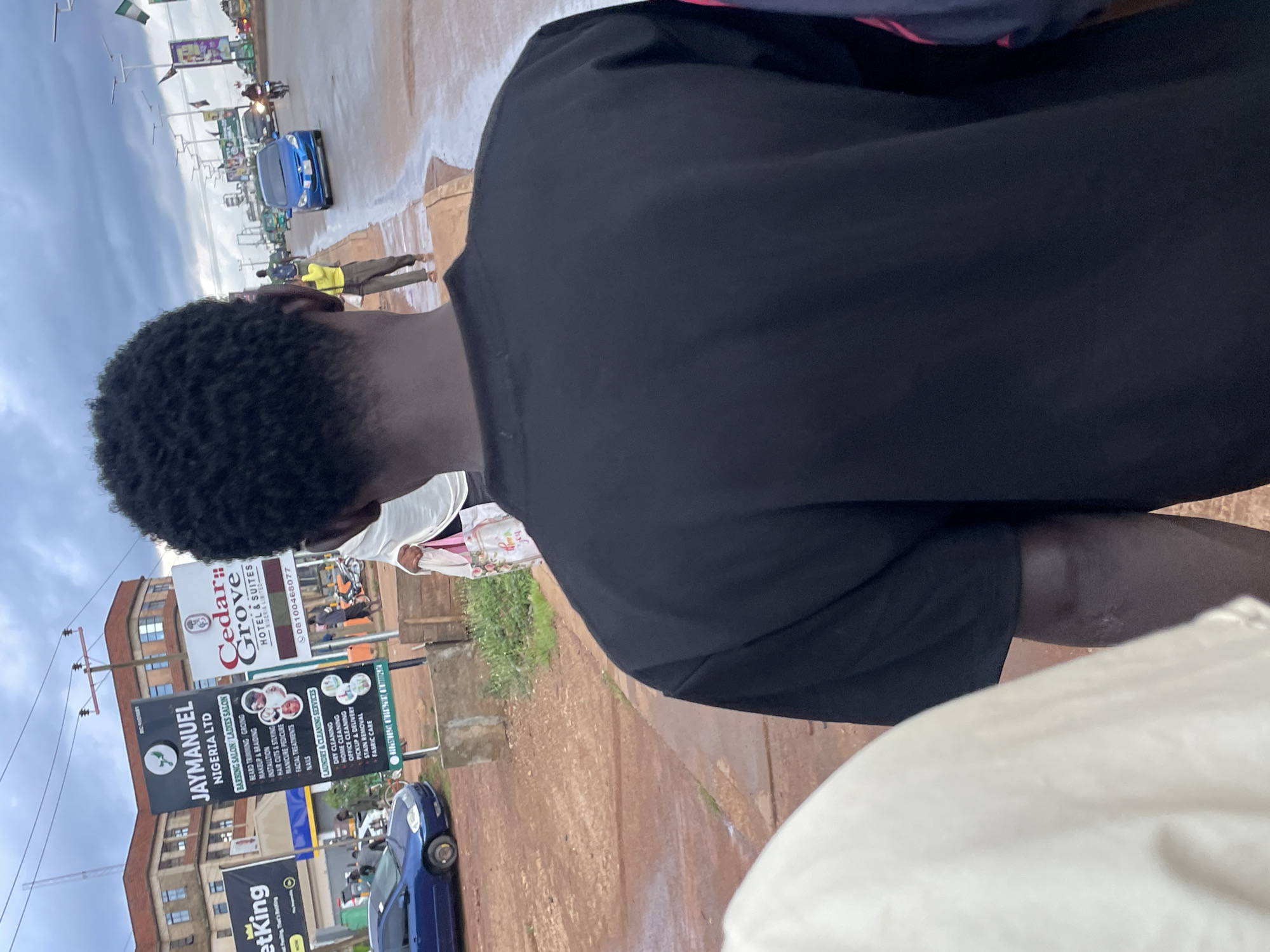 The image captures a street scene in Jos, Nigeria, under an overcast sky. In the foreground, the back of a person wearing a black t-shirt is visible. To the left, a sliver of another person in a light-colored garment is present. In the middle ground, several signs stand on poles. One prominent sign, for "JAYMANUEL NIGERIA LTD," lists services including "BARBING SALON / LADIES SALON," "BEARD TRIMMING & GROOMING," "MAKEUP & BRAIDING," "INSTALATION," "HAIR CUTS & STYLING," "MANICURE / PEDICURE," "FACIAL TREATMENTS," and "NAILS," as well as "LAUNDRY & CLEANING SERVICES" such as "DRY CLEANING," "HOME CLEANING," "OFFICE CLEANING," "PICKUP & DELIVERY," "STAIN REMOVAL," and "FABRIC CARE." Adjacent to this sign is another for "Cedar Grove HOTEL & SUITES," with a phone number.
Further down the street, vehicles are visible, including a blue car, a motorcycle, and what appears to be a commercial bus. People are walking along the roadside and on the street. The environment features a paved road, a sidewalk, and some greenery. The overall impression is of a bustling urban environment in Jos, Nigeria.