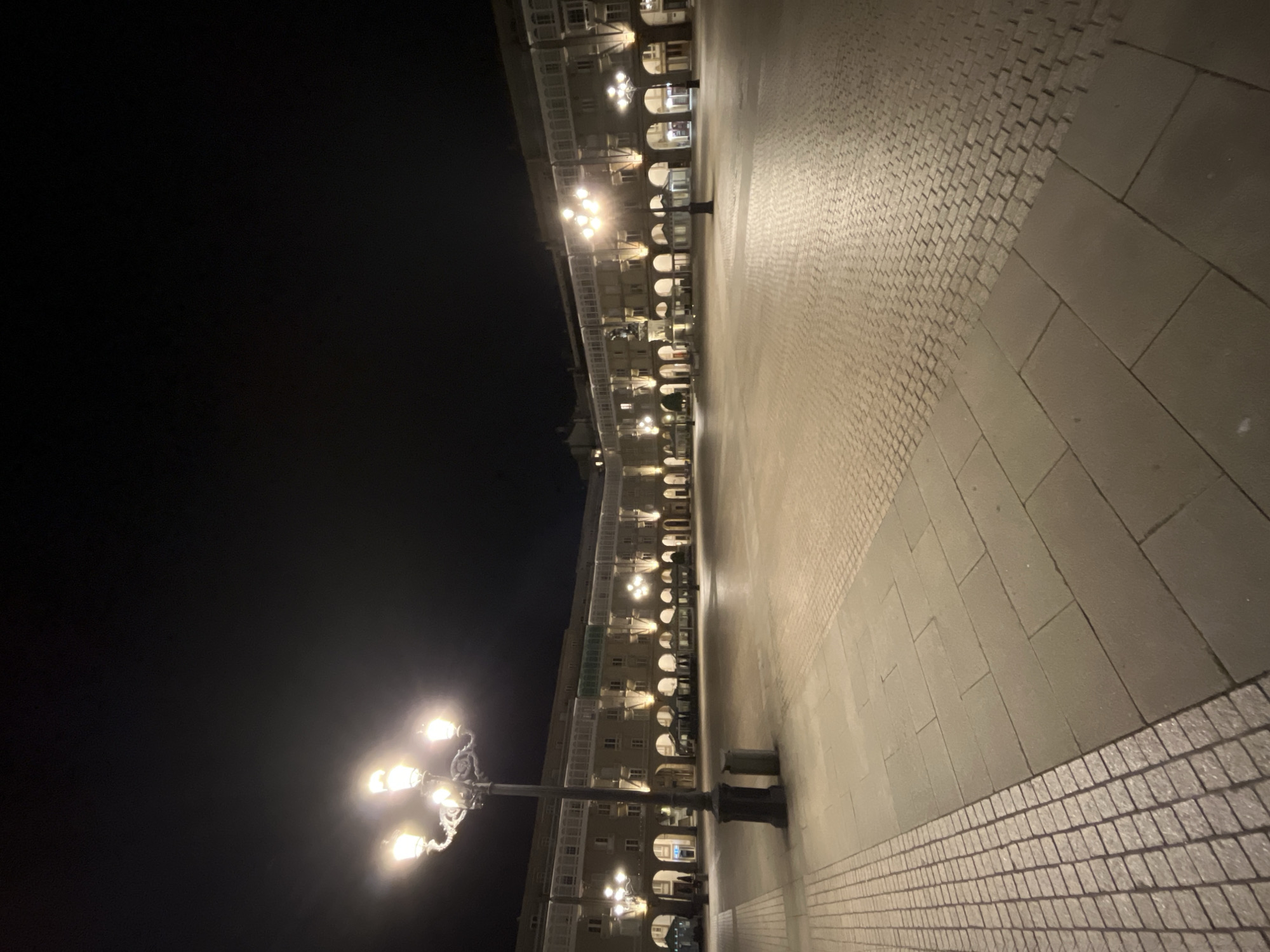 The image depicts the architecture of A Coruña, Spain, at night. A colonnade of buildings, illuminated from within and by external lamps, lines one side of a paved plaza. The pavement consists of a mix of large stone slabs and smaller cobblestones. A vintage-style street lamp with multiple bulbs stands in the foreground. The night sky above is dark. There is no visible human activity.