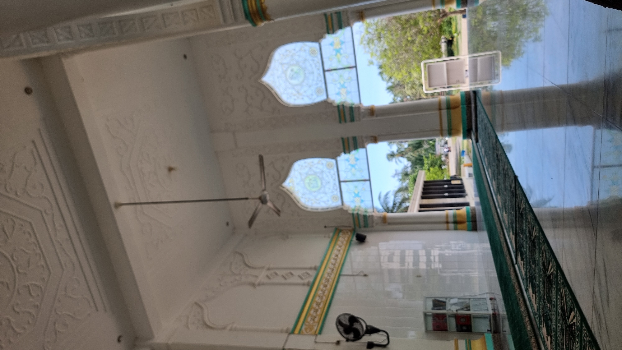 The image depicts the interior of a mosque with ornate white ceilings featuring geometric patterns. Two large, arched windows with colorful stained glass designs are visible, framing a view of palm trees and a clear blue sky. A ceiling fan is suspended from the center of the ceiling. The walls are white with decorative moldings in gold and green. A green carpet runs along the floor, which is highly polished and reflects the light and surroundings. To the left, a black oscillating fan is mounted on the wall, and a glass display cabinet containing books is positioned nearby. In the background, a section of a building with pillars and a dark facade is visible, along with more trees and what appears to be a road with a few people and vehicles. The scene suggests a well-maintained and architecturally detailed religious space. The location is identified as Lapang Timu, Indonesia.