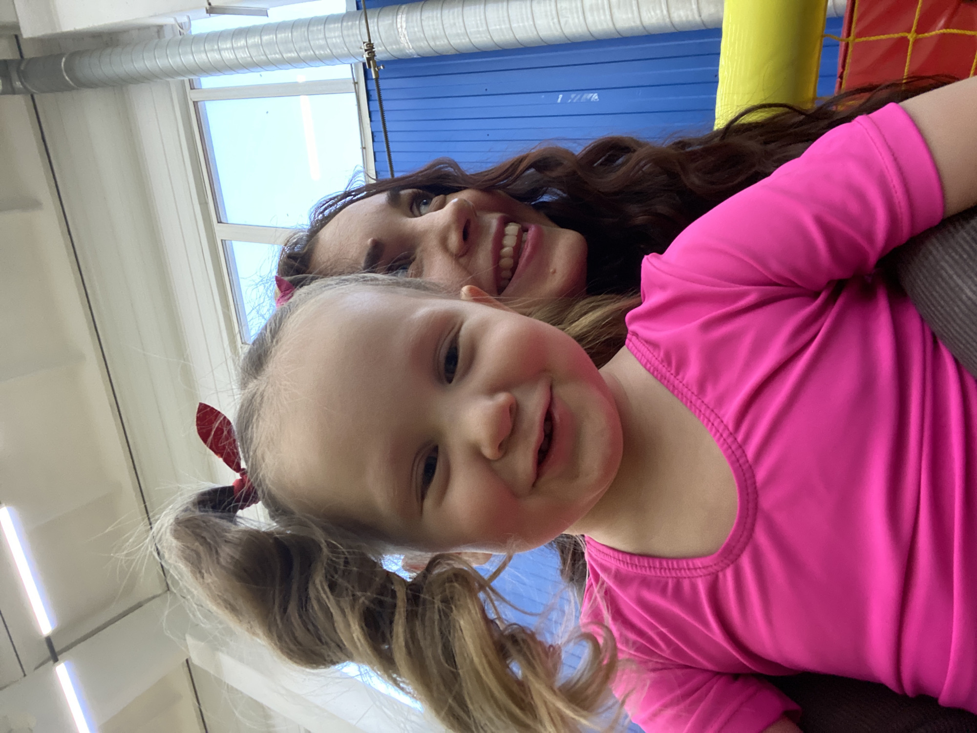 A mother and her young daughter are captured in a close-up selfie, likely taken at a children's play area. The location is implied as Chaikovsky, Russia. The daughter is in the foreground, wearing a bright pink shirt and with blonde hair in pigtails, each adorned with a red bow. Her mother is visible behind her, with long brown curly hair. Both are smiling at the camera. The background includes elements of the play area, such as a blue wall, a yellow tube, and possibly a padded structure. Natural light is coming in from windows, casting a brightness on the scene.