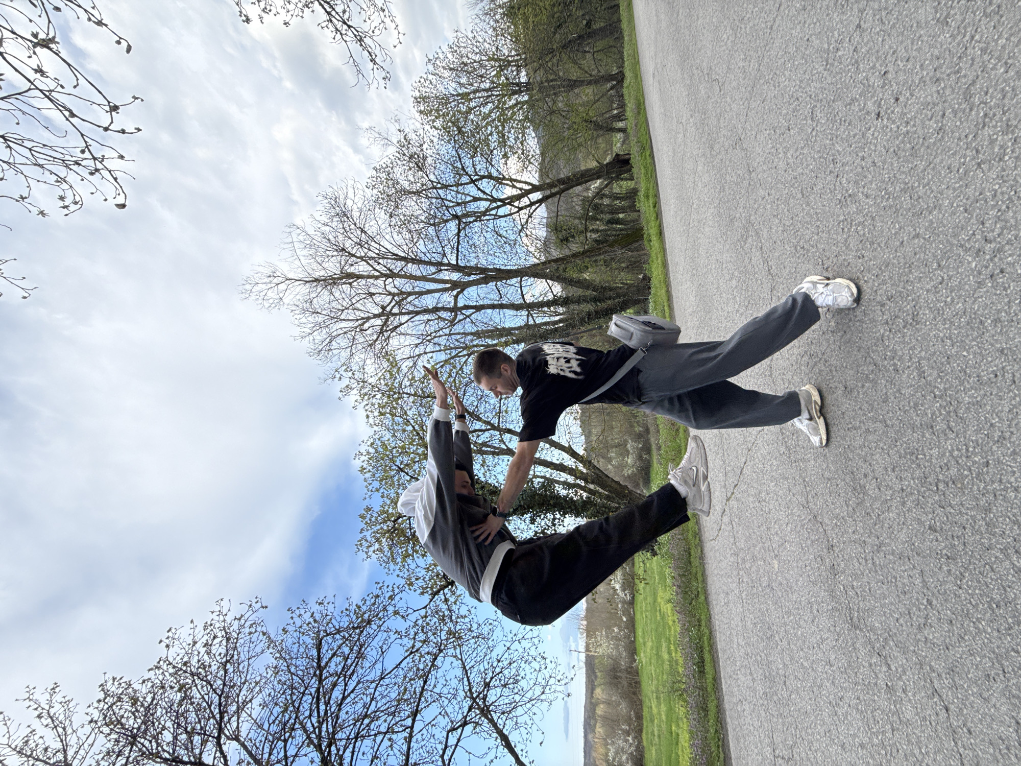 Two people are performing a physical stunt on an asphalt surface with green grassy areas on the side. One person is supporting the other with their arm, while the supported person is in a horizontal position with arms raised towards the sky. The background includes trees with bare branches and a partly cloudy sky. The scene appears to be outdoors in a park setting. The specific location in the city of Veliki Preslav, Bulgaria cannot be determined from the visual information provided.