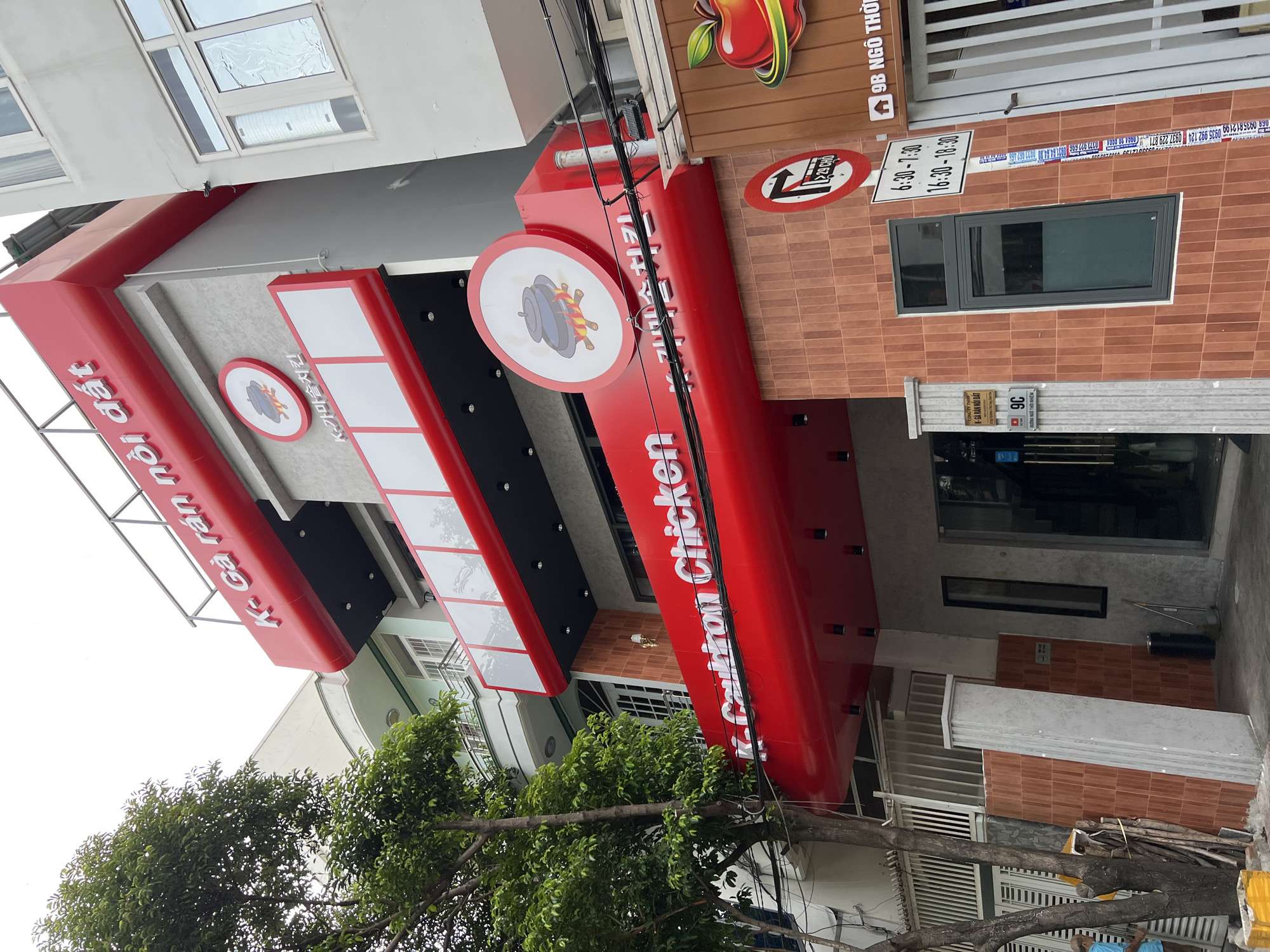 This is a full, eye-level outdoor shot of a modern, multi-story restaurant with a bright red facade. The restaurant appears to be a Korean fried chicken establishment, indicated by the "K- Ga rán nôi dat" signage in white lettering on the red exterior and Korean characters on a circular logo featuring a pot over flames. The building is narrow and tall, with multiple levels of windows and balconies. A large, leafy green tree is to the left of the building, partially obscuring a smaller adjacent building. There are street signs on the right side of the building, including one indicating "9B NGO THÔ" and another showing operating hours. There is a trash can and a broom near the entrance. No people are clearly visible in the image. The building is located in a densely populated urban area, as suggested by the close proximity of other buildings and overhead electrical wires. The overall impression is of a commercial street in a Vietnamese city.