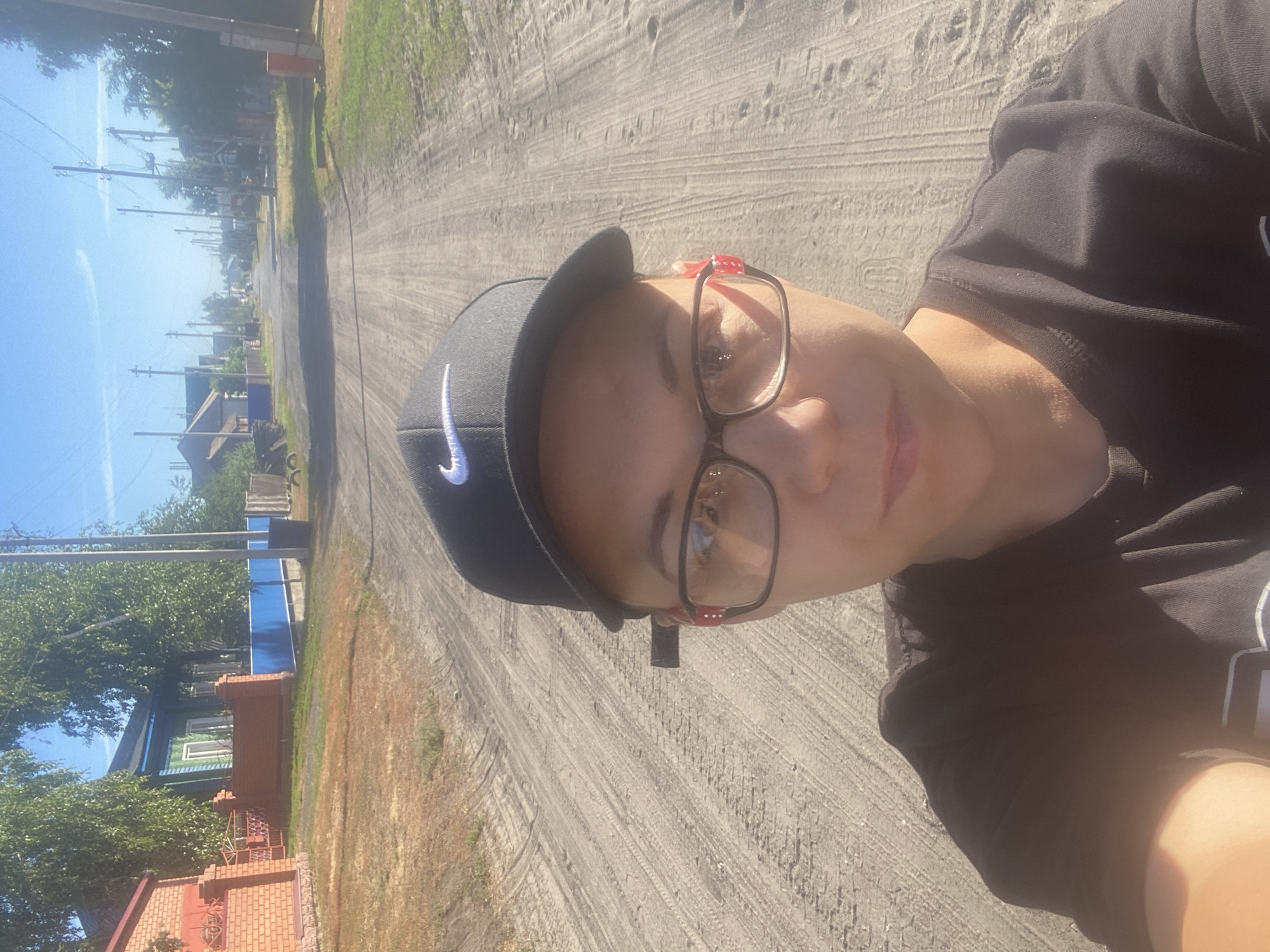 A selfie is taken from a low angle of a person wearing a black baseball cap with a white Nike logo, glasses with red accents on the temples, and a black t-shirt. The person is on a dirt road in what appears to be a village. In the background, there are houses with trees and telephone poles lining the road, suggesting a rural or suburban setting in Mamontovo, Russia. The road is unpaved and has tire tracks visible. The sky is clear and blue, indicating a sunny day.