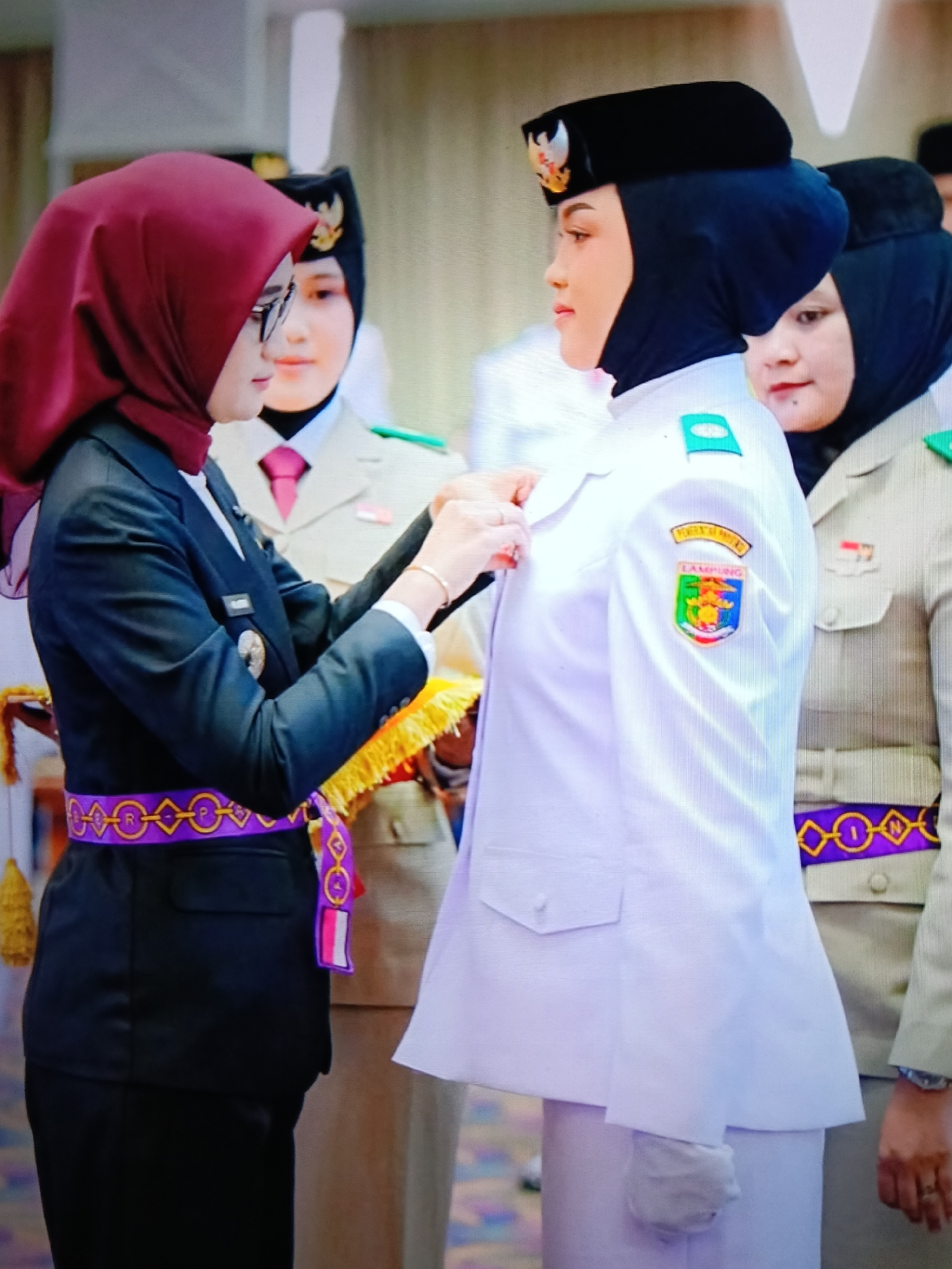 In Raha, Indonesia, two women in uniform are shown indoors. The woman on the left, wearing a dark suit and a maroon hijab, has glasses and is pinning a badge onto the uniform of the woman on the right. The woman on the right is wearing a white uniform, a black hijab and a black beret with an emblem. She has a badge on her left shoulder and a crest on her left chest. Another woman, also in uniform and wearing a hijab, is visible behind them on the right. Both women are adorned with sashes. The woman on the left has a purple and gold sash, while the woman behind her has a similar sash in purple and yellow. The background appears to be an indoor setting with muted colors.