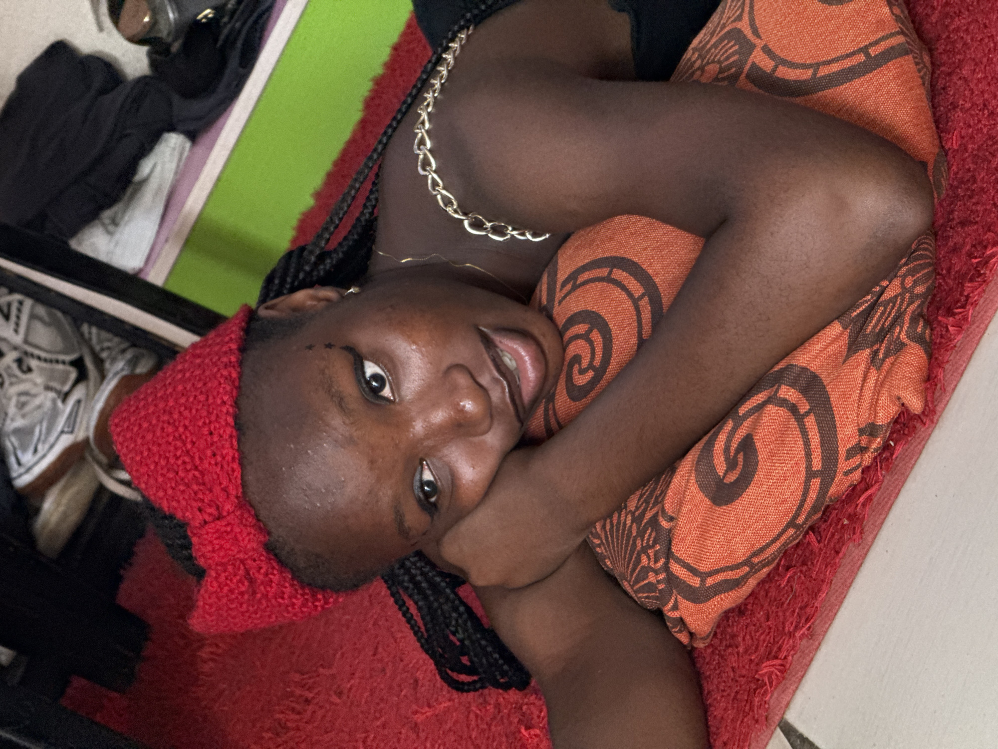 A young woman is lying on a red shag rug, hugging an orange pillow with a dark brown spiral pattern. She is facing the camera with her head tilted. The woman has dark skin, braided hair, and is wearing a red knit headband with a bow. She has a gold chain necklace and a thinner gold necklace. In the background, there is a green and purple wall, as well as a partial view of shoes and clothing. The location is in Abuja, Nigeria.