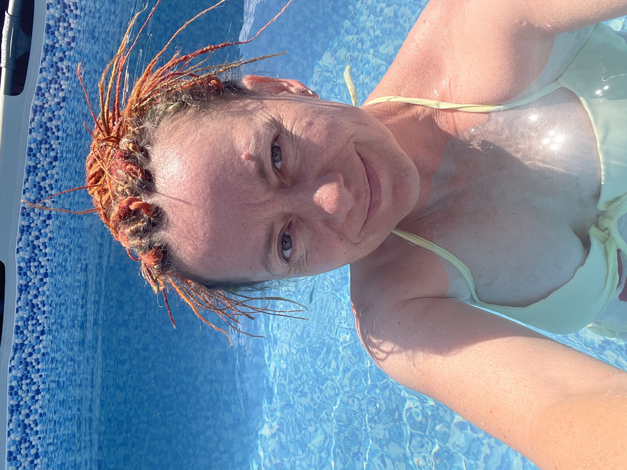 A woman with auburn dreadlocks smiles at the camera while swimming in a bright blue pool. The pool has a tiled border with blue and white patterns. The water is clear and rippling. She is wearing a light yellow bikini top. The setting appears to be a sunny day, likely outdoors. No specific location in Orenburg, Russia, can be identified from the image.