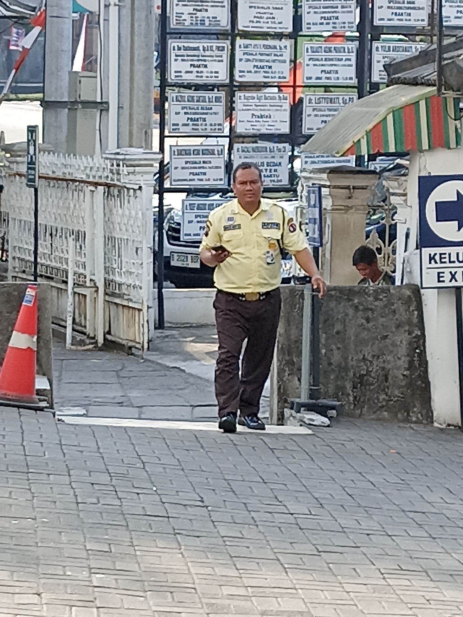 A security guard in a light yellow uniform with patches on the sleeves and a nametag walks on a paved walkway. He is holding a mobile phone in his right hand. Behind him, a young man in a camouflage shirt is visible behind a concrete barrier, looking down. Several signs with text in Indonesian are mounted on a pole to the left of the security guard. To the left of the walkway, a white metal fence runs along the edge of the path. A red traffic cone is positioned near the fence. In the background, a white vehicle is parked, and buildings with multiple windows are visible. The scene appears to be a public area in Beji, Indonesia, likely near a medical facility given the signs.