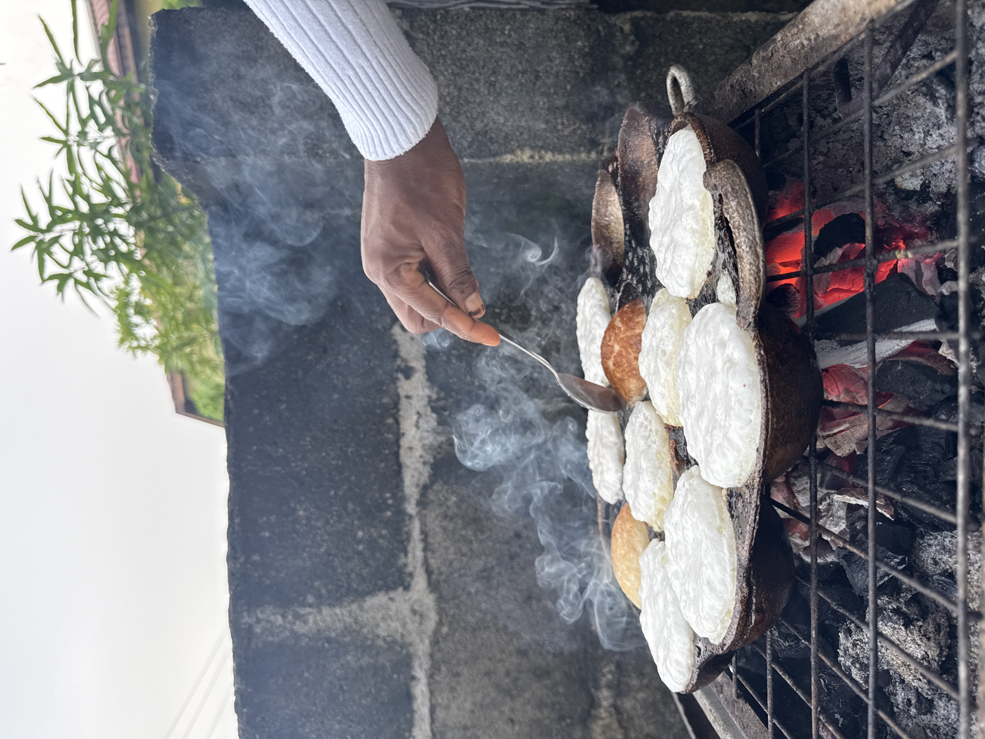 In Ado, Nigeria, a person with dark skin and wearing a white sweater is using a metal spoon to cook a local delicacy in a traditional grill. The grill is placed over hot coals, producing smoke. The delicacy consists of small, round cakes, some are white and some are light brown. The grill itself is made of a series of cast iron pans forming a complete circle. The pans are placed on a mesh rack that is above hot coals. The scene appears to be in a backyard or outdoor cooking area. A concrete wall is visible, as well as some green foliage and clear sky.