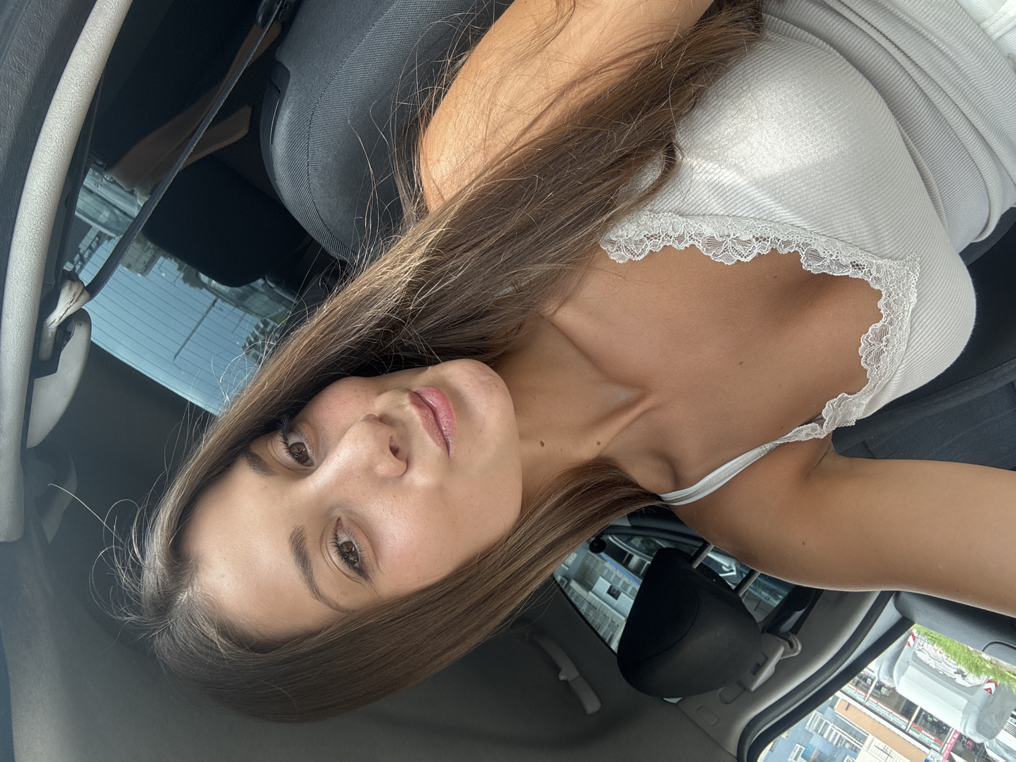A young woman with long brown hair is in the driver's seat of a car. She is wearing a white tank top with lace trim. The car is likely parked based on the view through the window. Reflected in the car's side mirror are buildings typical of an urban environment, possibly located in Burgas, Bulgaria. A seatbelt is visible across the seat.
