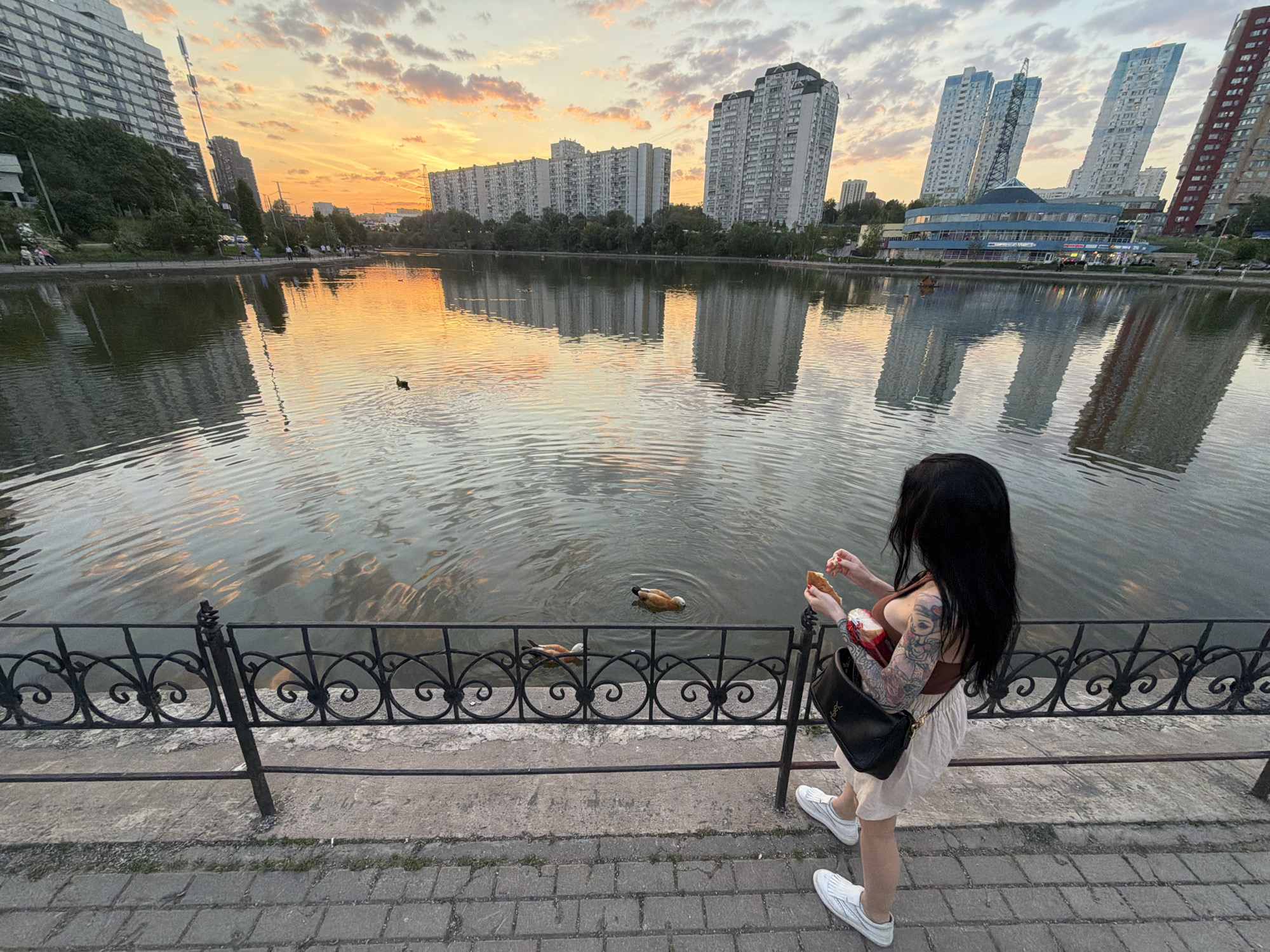 The photo depicts a woman standing next to a wrought iron fence beside a body of water, likely a pond or lake. She is facing the water and holding what appears to be food in her hands, possibly feeding ducks that are swimming in the water. The woman has dark hair and is wearing a tank top, a skirt, and sneakers. She has tattoos on her right arm and a black purse hangs from her left shoulder. The lake reflects the buildings on the far side, as well as the sky and the buildings in the background. In the background are buildings and trees. The sky is visible above the buildings and is a mix of colors including yellow, orange, and white, suggesting it is either sunset or sunrise. The photo was taken in Moscow, Russia.