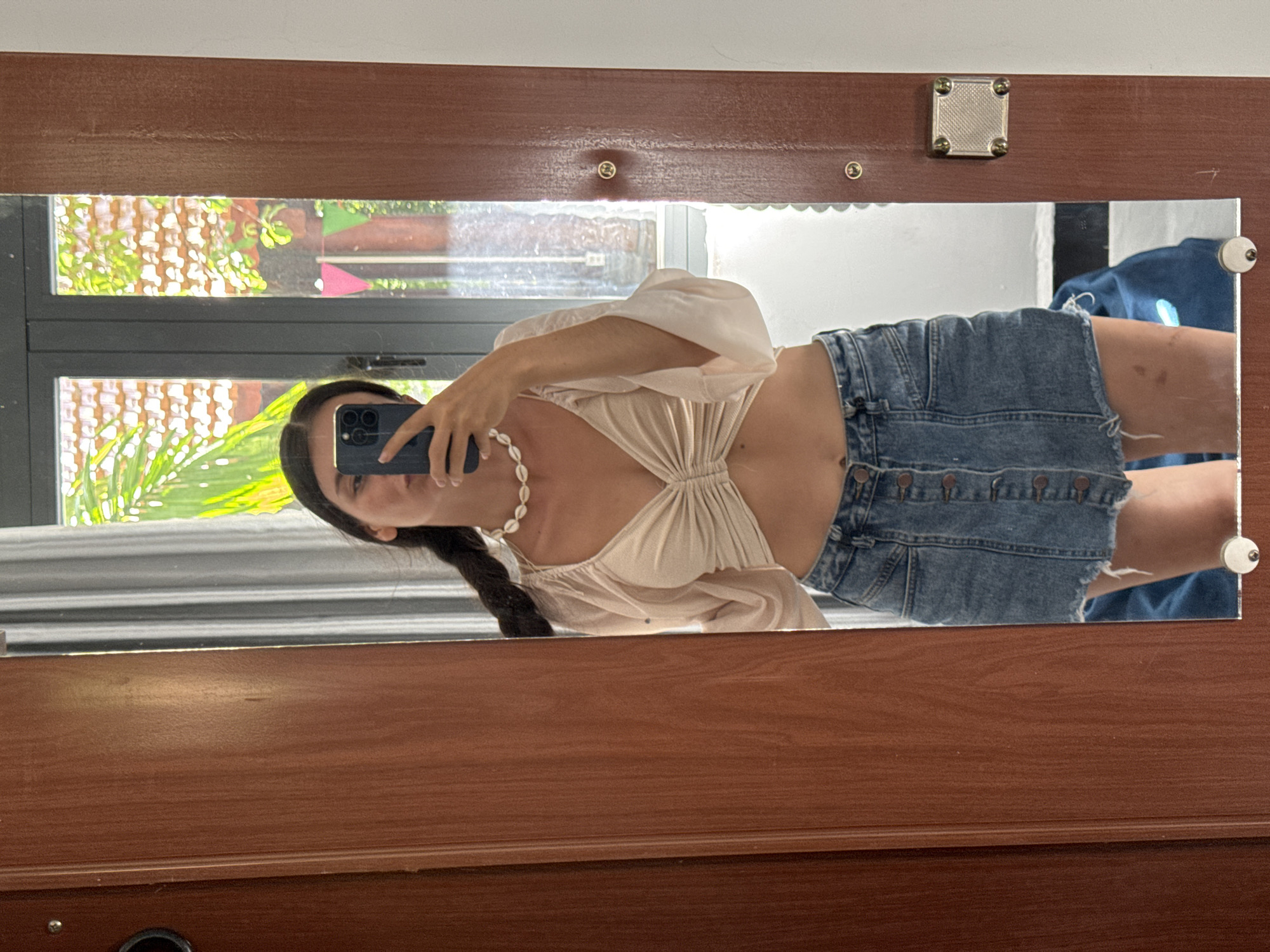 A woman is taking a mirror selfie in Mũi Né, Vietnam. She is wearing a cream-colored crop top with ruching in the front and a denim mini skirt. Around her neck, she has a white shell necklace. The background visible in the mirror includes a window with a view of lush greenery, red-tiled roofs, and some colorful triangular flags. To the left of the frame, a wooden door is visible, and to the right, the white wall meets the wooden doorframe. The overall setting appears to be an indoor space, possibly a hotel room or a home, with a view of the outdoors through the window.