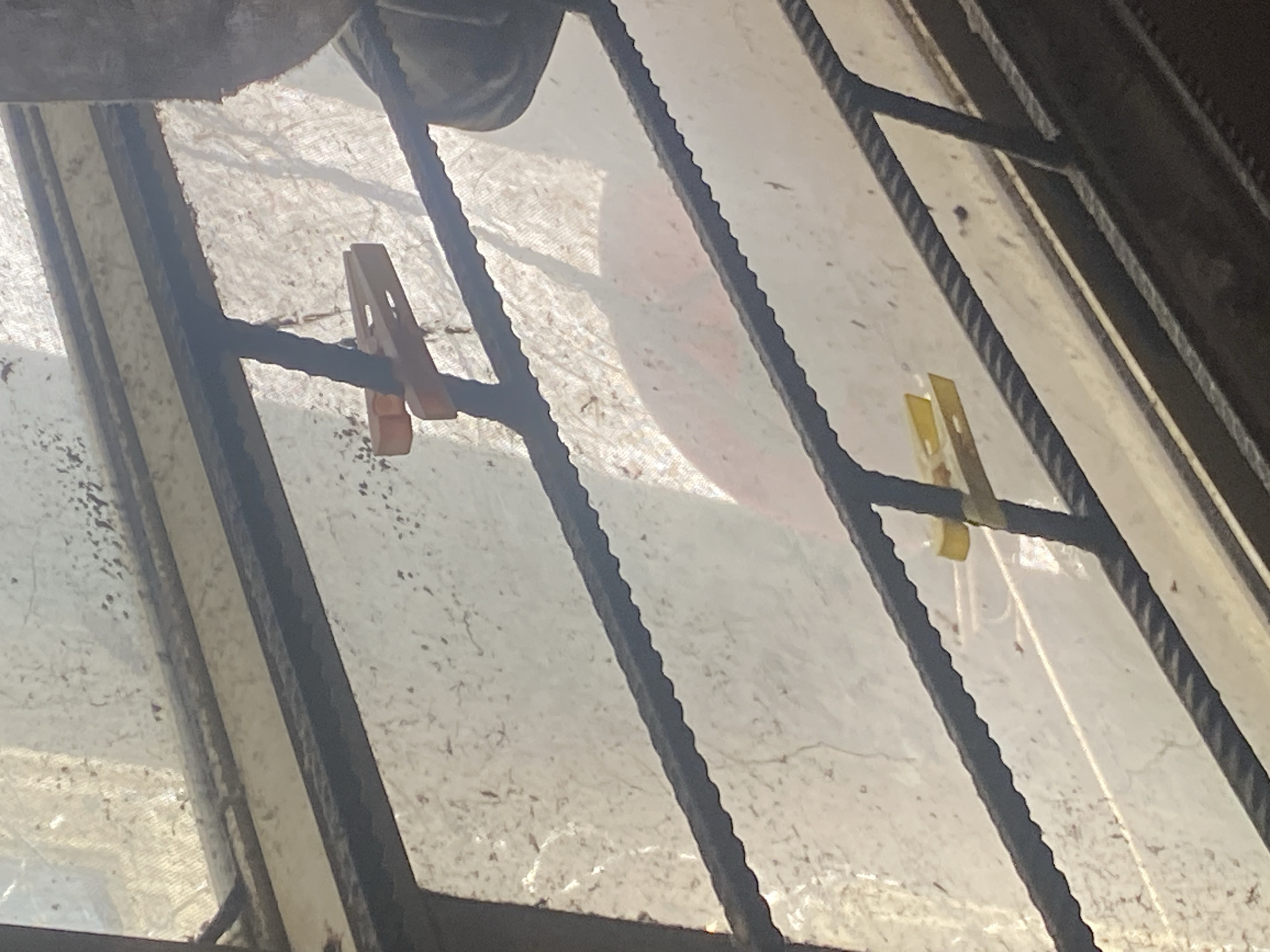 The image presents a close-up view through a grimy, translucent barrier, likely a window or opening covered by a plastic sheet or tarp. Dark, corrugated metal bars, appearing weathered and possibly rusty, crisscross the frame, suggesting a security grille or reinforcement. Bright daylight filters through the translucent material, illuminating its textured and somewhat dirty surface.
Two plastic clothespins are clipped onto the metal bars. On the left, an orange clothespin secures the translucent covering to a horizontal bar. On the right, a yellow clothespin is attached to another bar, with a thin white string or cord hanging loosely from it. Behind the translucent barrier, indistinct shapes and shadows are visible, including a soft, reddish-orange blur in the upper right.
The scene captures a utilitarian setting during daytime, under bright sunlight, possibly in Ikorodu, Nigeria. The use of clothespins and the condition of the materials imply a practical or makeshift arrangement. No specific activity or event is visible, nor is any text present.