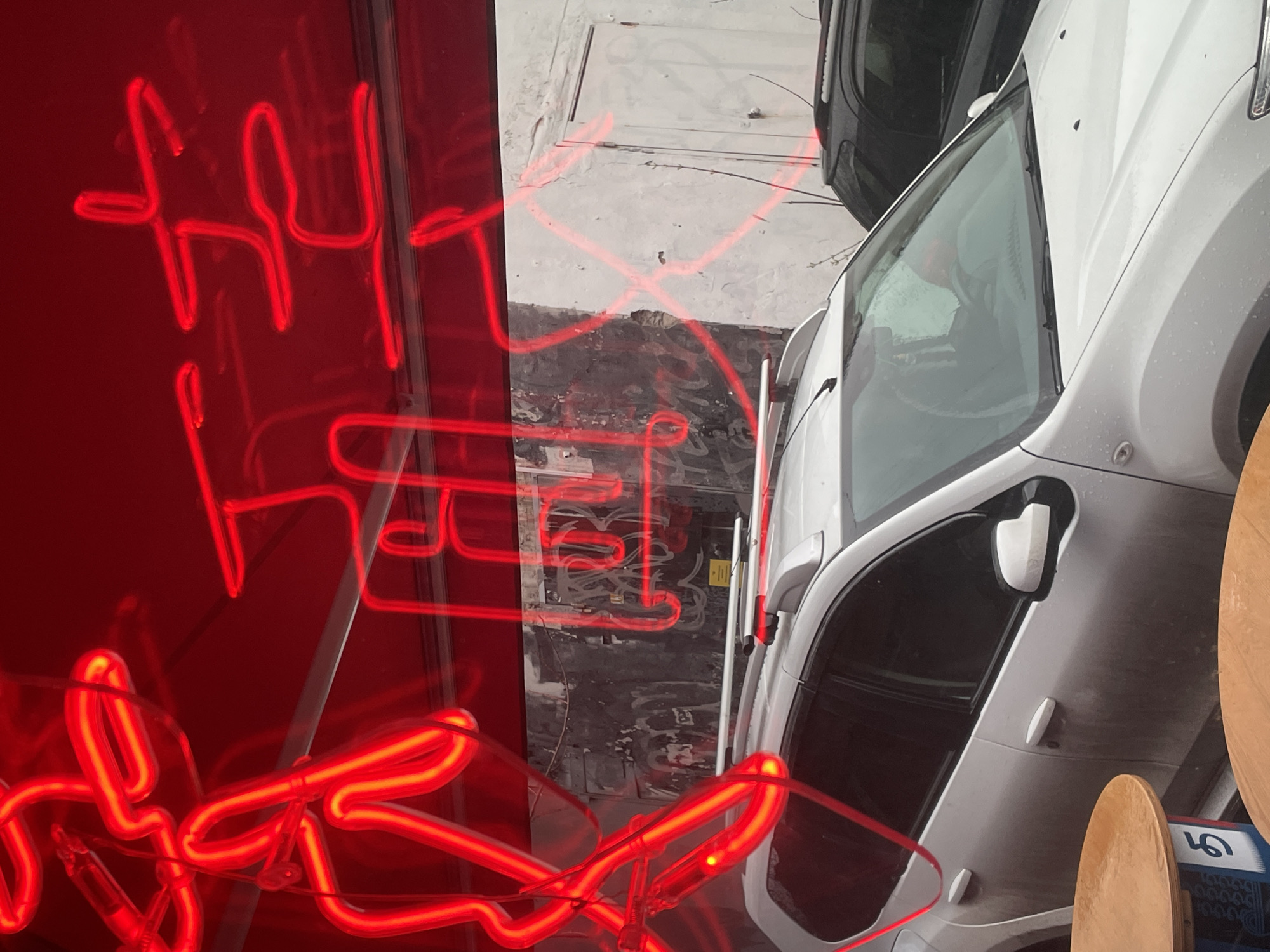 The image depicts a street scene in Perm, Russia, viewed from above. A white vehicle is parked alongside a building with a reflective red sign. Red neon lights create reflections on the vehicle, the sidewalk, and the building. A black vehicle is partially visible in the upper right corner of the frame. The sidewalk appears wet. The letters of the red sign are visible in the reflections, but the full text is obscured. A wooden object, possibly a board, is positioned near the right side of the white vehicle.