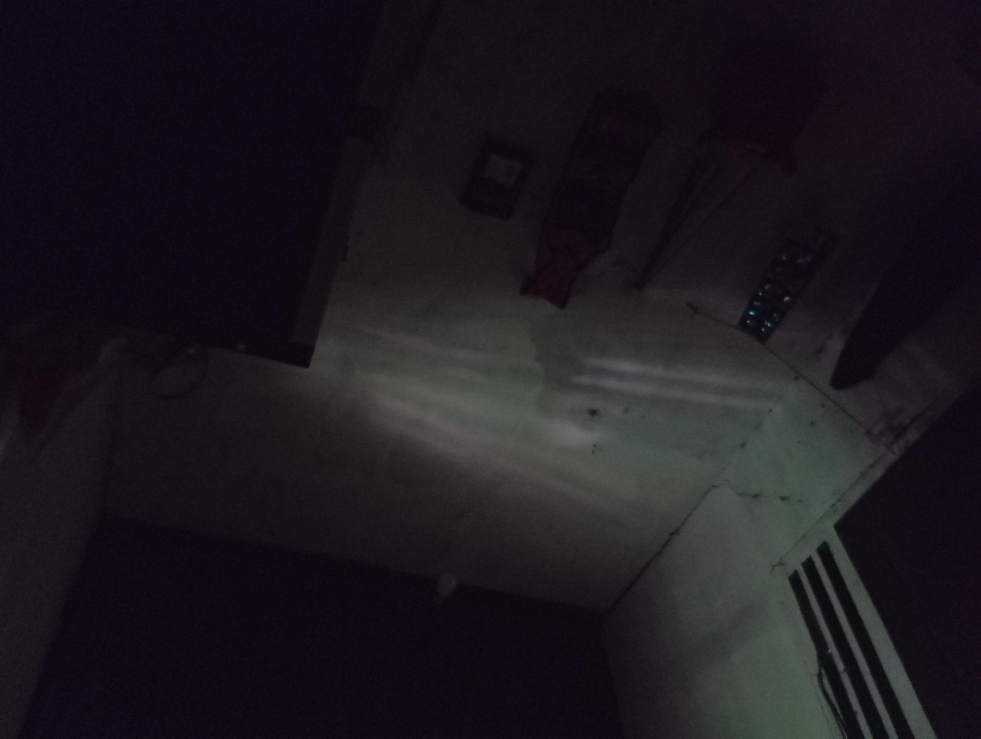The image depicts a dimly lit, seemingly empty room. The walls appear to be concrete or a similar material, with visible cracks and scuff marks. In the foreground, a dark, amorphous shape suggests the presence of an object or furniture, possibly a table or a large box, which is mostly obscured by shadow. To the left of this dark object, a rack with several small, illuminated, rectangular objects is visible, possibly batteries or electronic components. Further to the left, there's a suggestion of a black, vertical surface with three parallel white stripes. In the mid-ground, a red, bag-like object with straps is lying on the floor, and next to it is a small, dark rectangular object with a lighter rectangular area on its surface, perhaps a small electronic device or a label. The floor itself is stained and appears to be concrete, with streaks of light reflecting off its surface, indicating a light source outside the frame. The ceiling is largely obscured by darkness, though a faint, white circular object is visible in the upper right portion of the frame. The overall impression is one of disuse or a storage area. The location of Rejosari, Indonesia is not discernible from the visual information provided in the image.