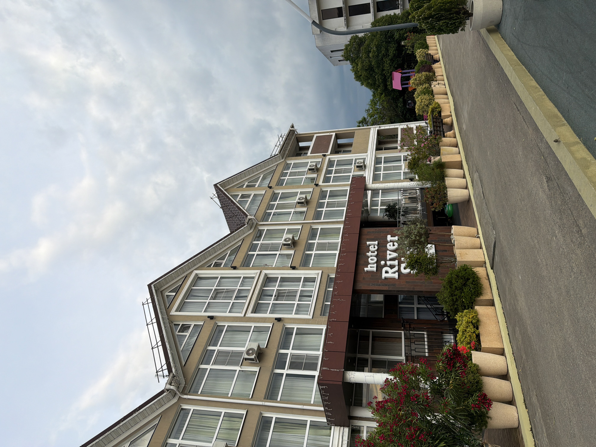 This image shows the exterior of the Hotel River in Sochi, Russia. The hotel is a multi-story building with a distinctive sloped roofline and numerous windows. The facade is a light brown color, with dark brown accents around the entrance. The hotel's name, "hotel River", is visible in white lettering on a dark brick section near the entrance.
In front of the hotel, there is a walkway lined with large, beige planters containing various green shrubs and some flowering plants with red blooms. A paved path runs alongside the planters, separating them from the hotel. In the background, behind the hotel, there are trees and a glimpse of another building. A pink playground structure is visible among the trees, suggesting a family-friendly environment.
The sky is overcast with scattered clouds, indicating a cloudy day. The lighting is diffused, suggesting it is daytime, possibly late morning or afternoon. The overall scene depicts a hotel entrance area designed with a focus on landscaping and guest welcome. There are no people visible in the image. The image is captured from a low angle, looking up towards the hotel.