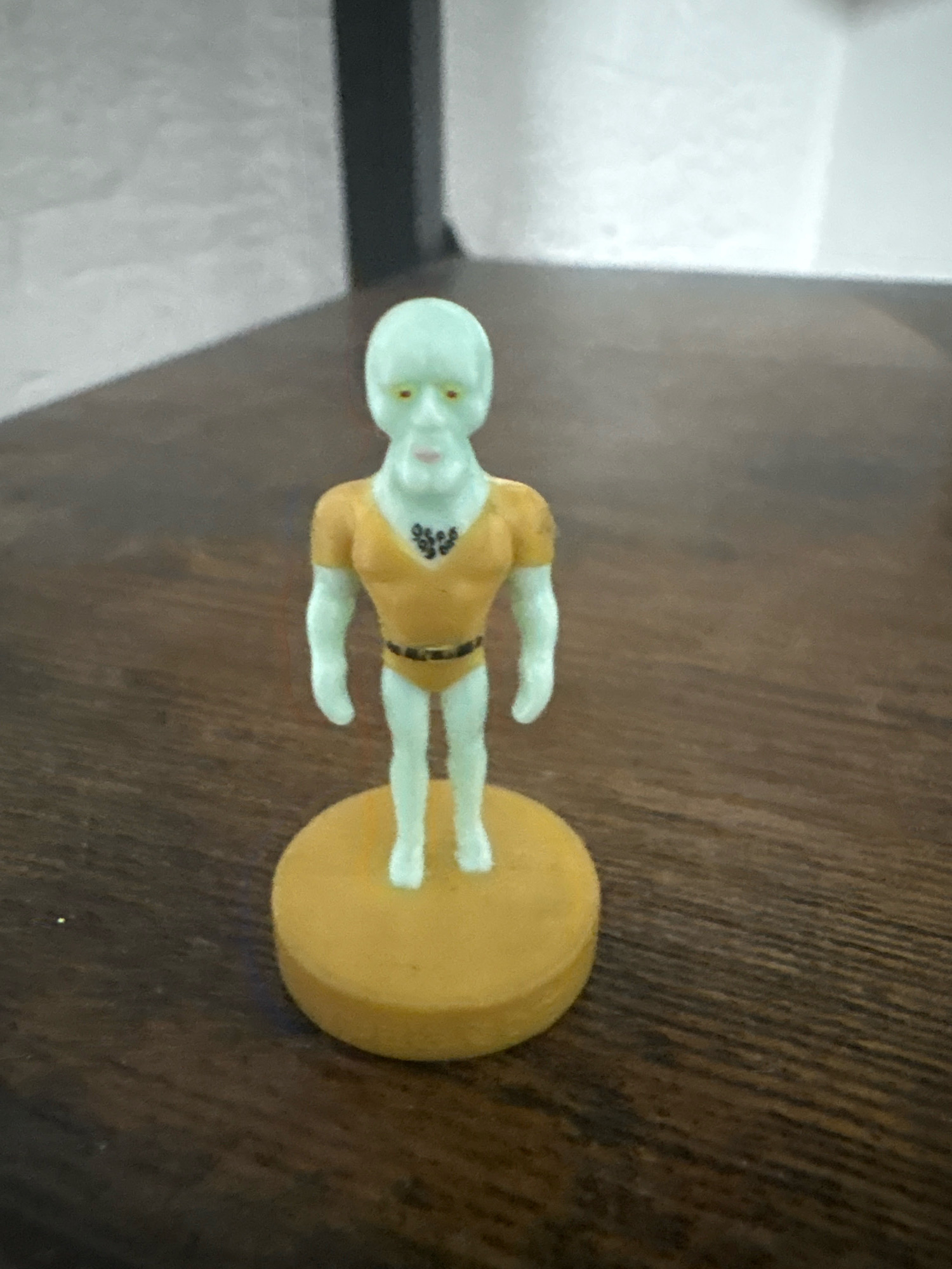 A light green, anthropomorphic figurine of a character resembling Squidward Tentacles from SpongeBob SquarePants stands upright on a round, yellow base. The figurine has a bald, pale green head with yellow eyes and a small mouth. It is wearing a short-sleeved, orange-yellow shirt with black detailing on the chest, a black belt, and orange-yellow bottoms. The figurine's arms are extended slightly downwards at its sides. The yellow base is circular and flat. The figurine and base are situated on a dark brown, wooden surface that exhibits a wood grain pattern. In the background, a blurred, light-colored wall with vertical lines is visible, casting a shadow on the wooden surface. The scene appears to be indoors.