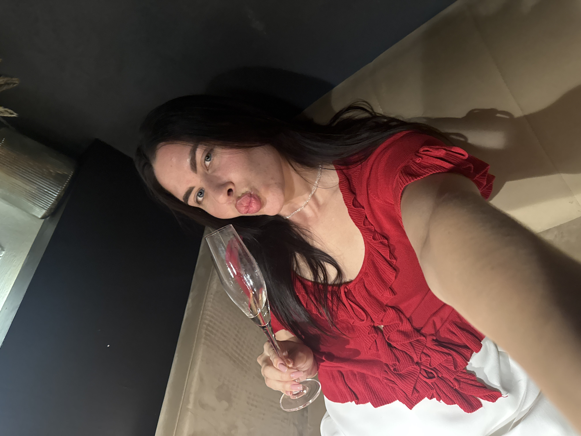 A young woman with dark hair and blue eyes is sitting on a couch, holding a champagne flute. She is wearing a red, ruffled top and a necklace. She is puckering her lips as if to kiss the camera. The background is a dark wall. The image is a selfie, taken indoors. The location in the city of Chaikovsky, Russia, is not discernible from the image.