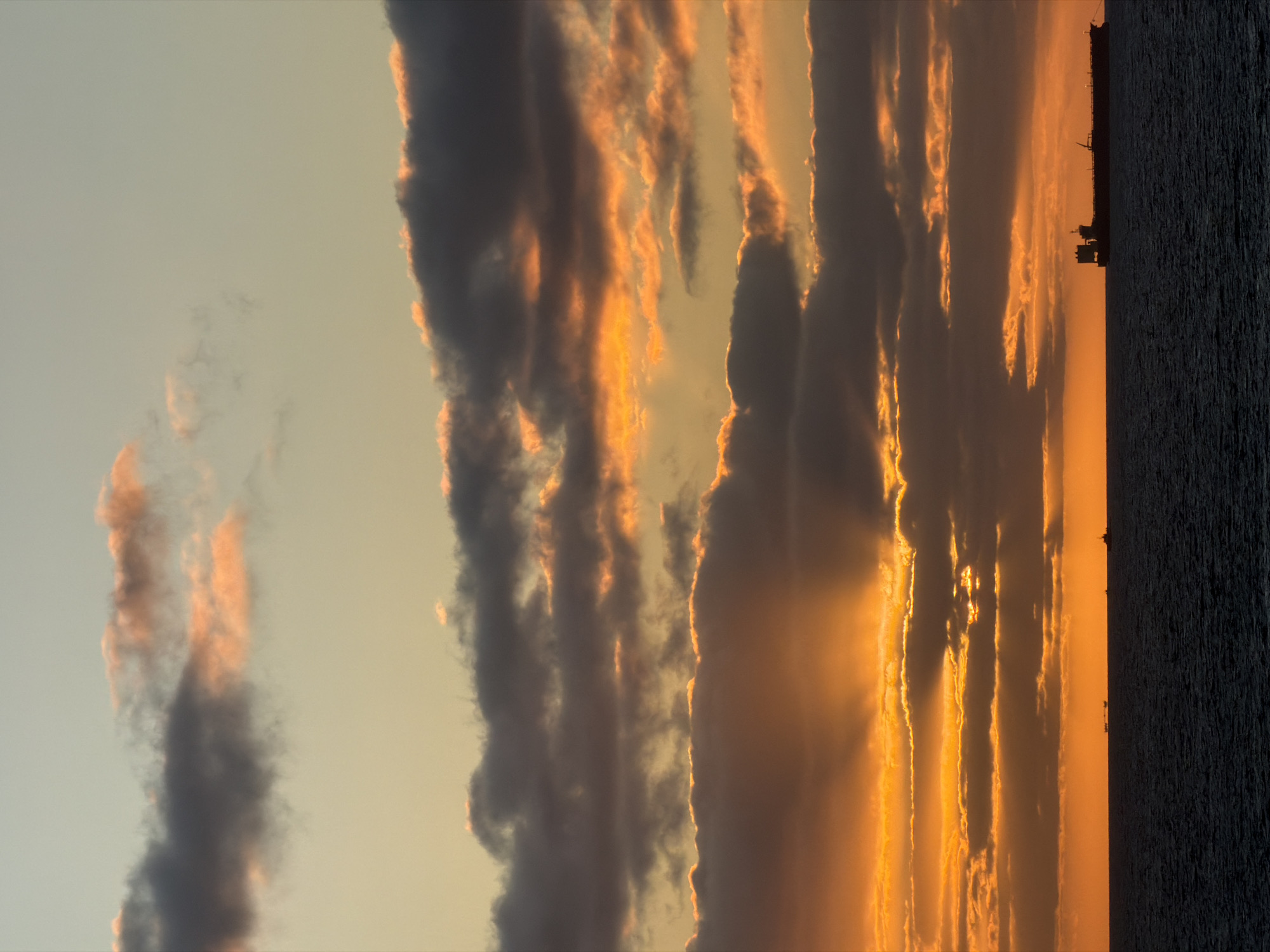 Here is a detailed description of the image:
The image depicts a vibrant sunset over the ocean, possibly in a coastal area. The sky is filled with clouds, many of which are illuminated with a golden-orange hue from the setting sun. Some clouds are darker, creating a dramatic contrast. Rays of sunlight peek through the clouds, adding to the scene's intensity. A dark silhouette of a ship is visible on the horizon against the bright sunset. The ocean is a dark, textured mass, contrasting with the luminous sky. The overall impression is one of natural beauty and serenity, tinged with a dramatic sense of light and shadow.