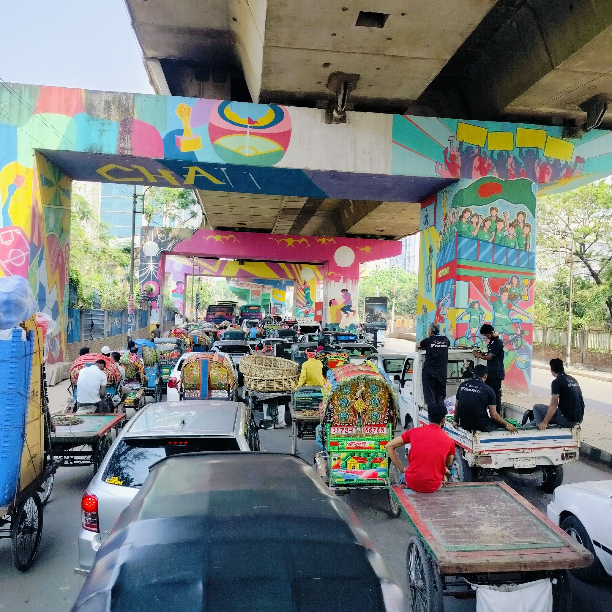 The image shows a congested street scene in Dhaka, Bangladesh, under an overpass adorned with colorful murals. The murals depict various scenes, including people holding signs, a soccer team with the word "BANGLADESH," and abstract designs. The traffic consists of a mix of cars and rickshaws, many decorated with elaborate designs and paintings. A group of men wearing "DAIKIN" branded shirts are standing on a truck and working on something attached to one of the overpass pillars. The sky is overcast, and the light suggests it is daytime. The license plates on the vehicles are visible, revealing details specific to Bangladesh.