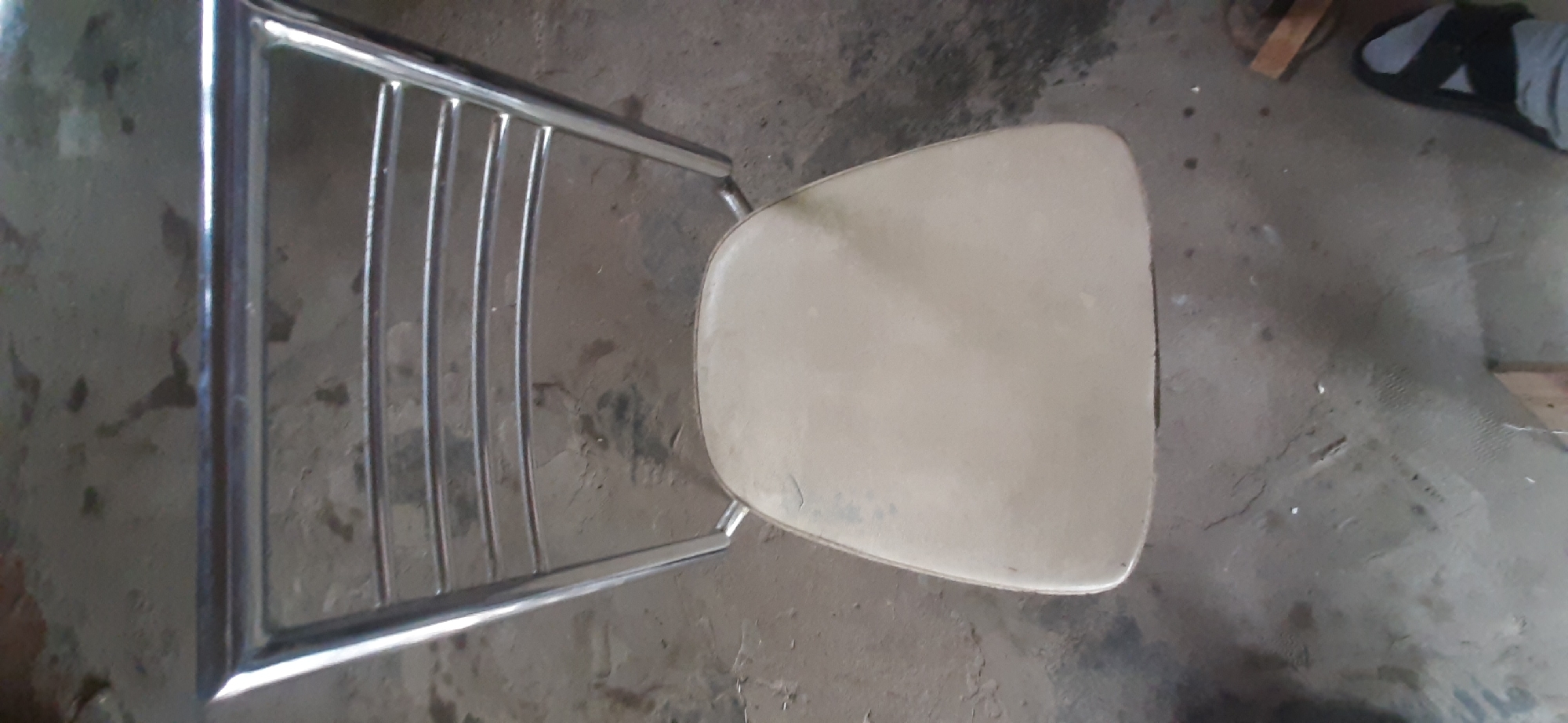 The image shows a high-angle shot of a metal-framed chair with a beige cushioned seat and back. The chair is positioned on a concrete floor, which appears to be indoors. The background is indistinct, showing areas of shadow and light. There are no people visible in the immediate vicinity of the chair. The overall impression is one of an ordinary, utilitarian chair in a somewhat plain setting. There is no information available about the location within the city of Yessentukskaya, Russia, as the image itself does not provide any contextual clues to support this claim.